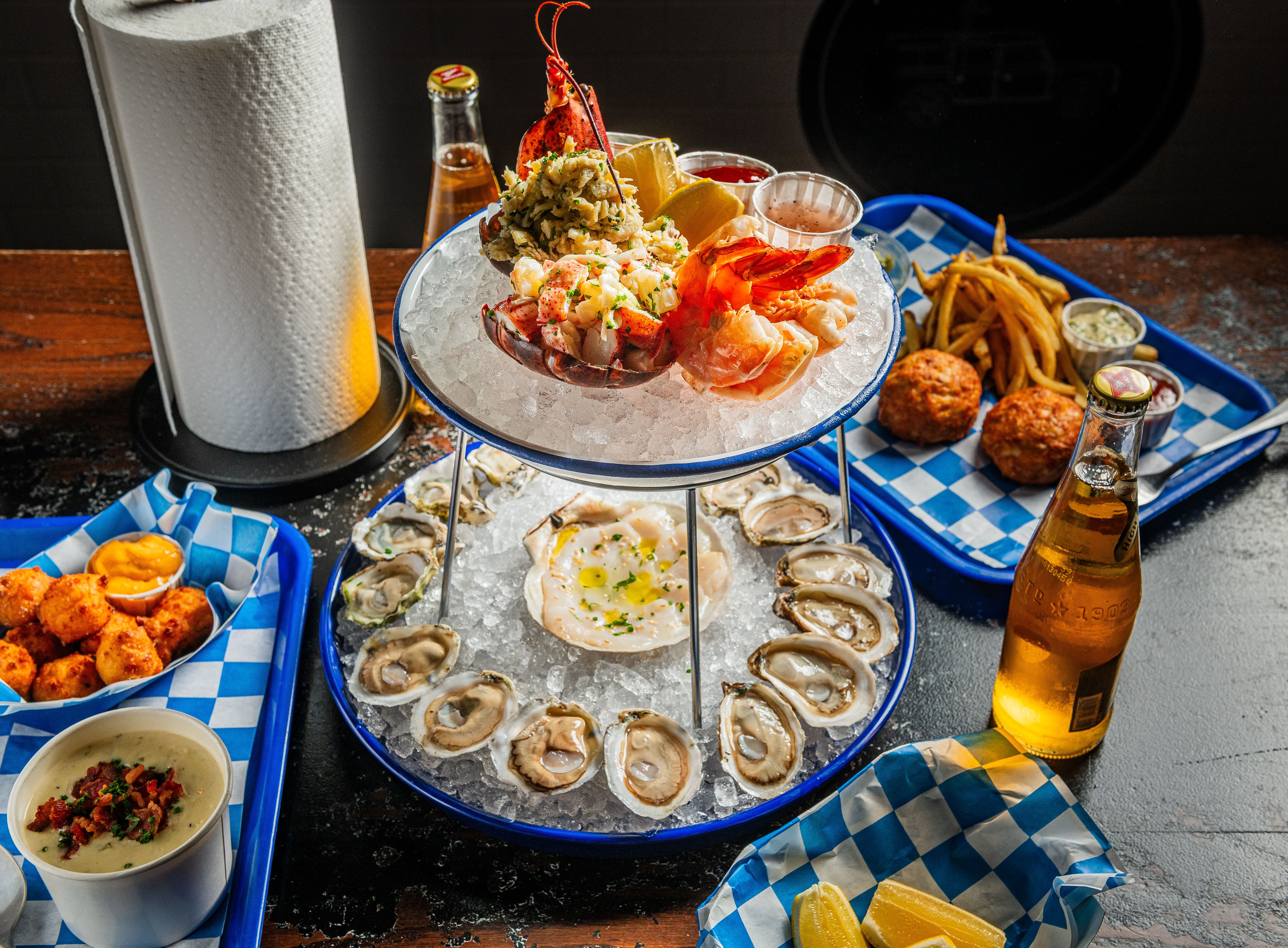 Two-tier seafood platter with oysters, shrimp, lobster on ice, lemon wedges, sauces, crab cakes, fries, cheese bites with dip, bottled drinks, and a paper towel roll on a dark table.