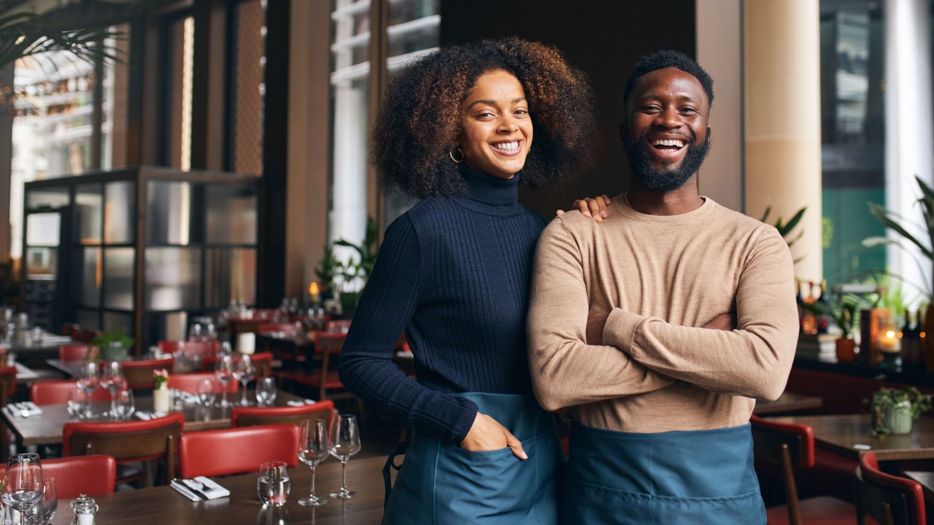 Smiling male and female restaurant staff in blue aprons standing inside a warmly lit, elegant dining area with red chairs and set tables.