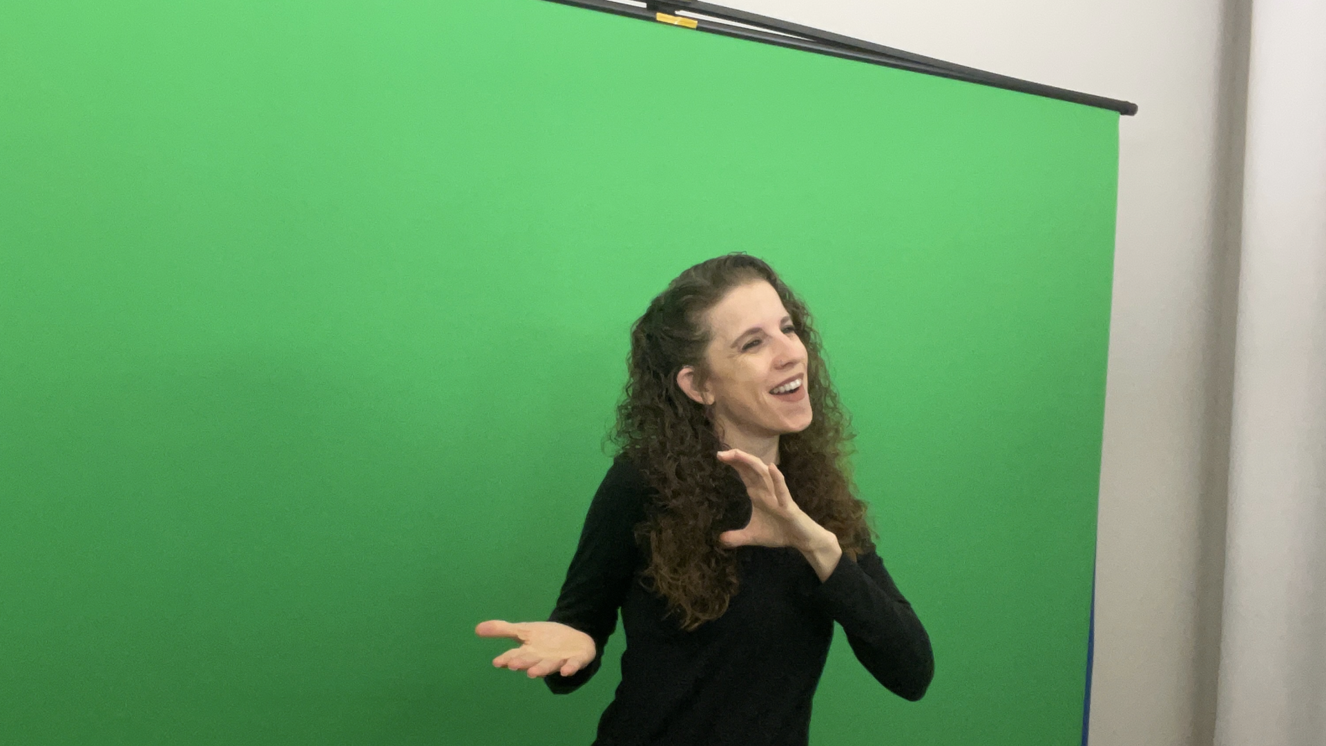 Woman with curly hair in a black long-sleeve top stands in front of a green screen, smiling and gesturing with her hands as if explaining something.
