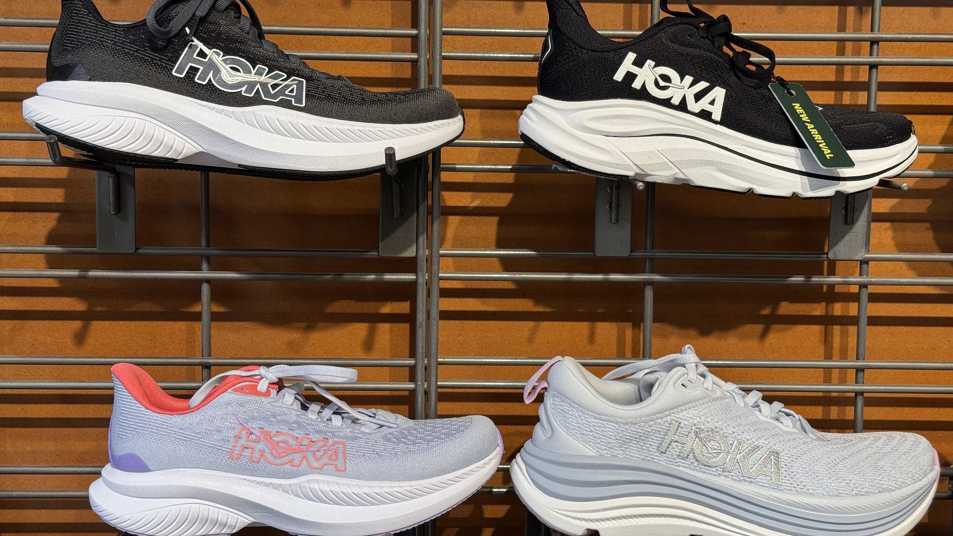 Four pairs of HOKA running shoes displayed on a metal grid wall; top two pairs are black with white soles, bottom two are light gray with pink and blue accents respectively.