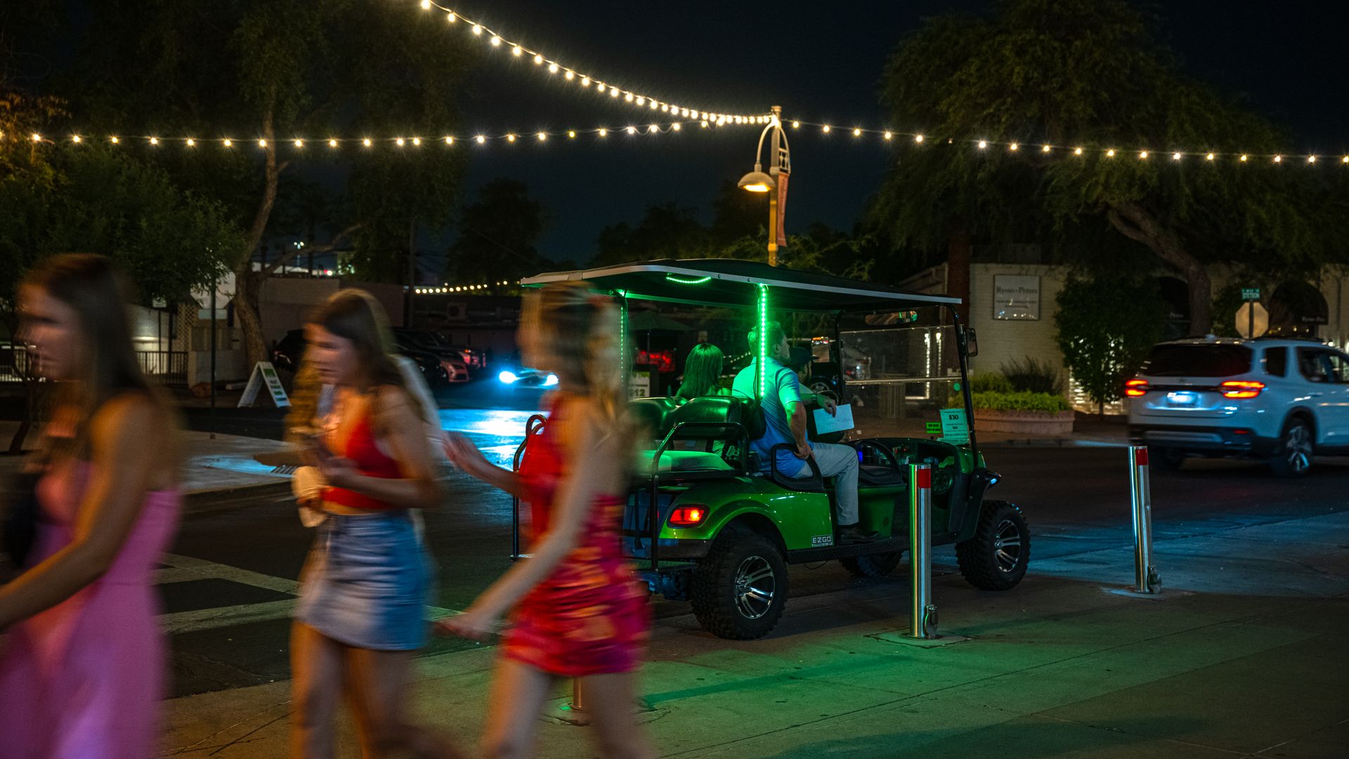 Three girls walking, gold cart in the background and string lights overhead and brightly lit in the dark sky.