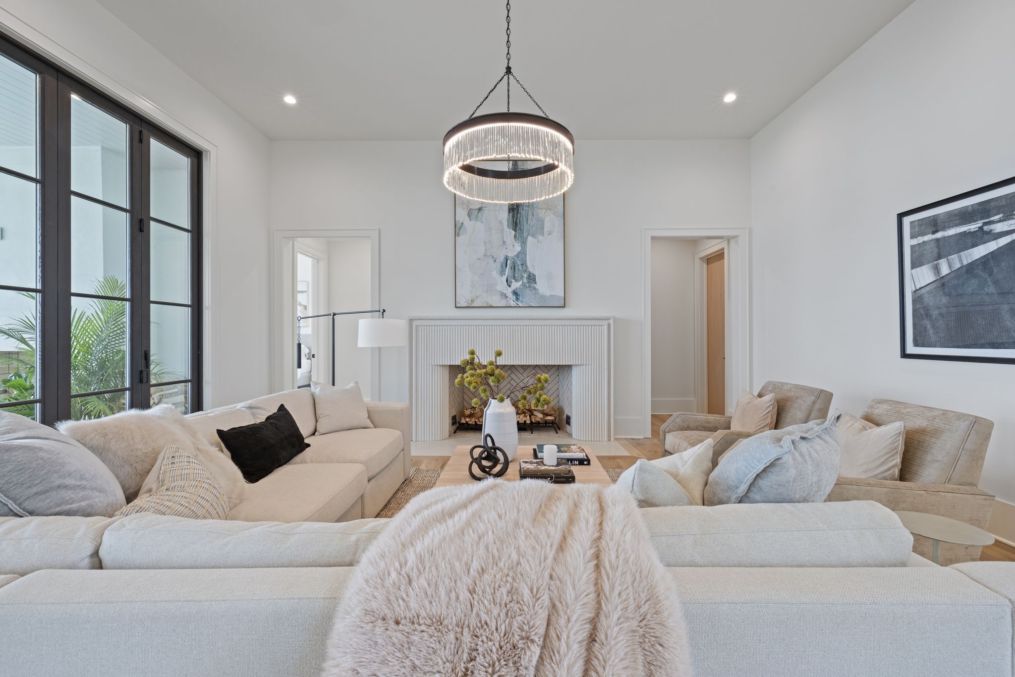 Bright living room with beige sofas and armchairs, black and white throw pillows, a modern chandelier, a white fireplace with abstract art above, and large windows with black frames.