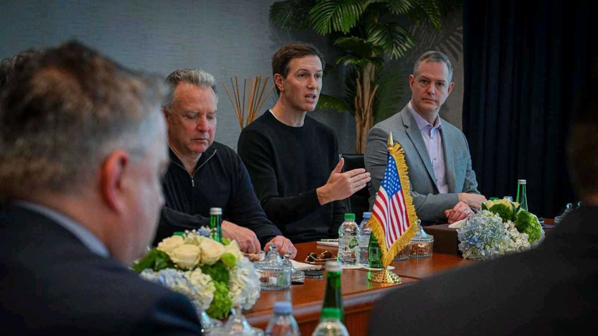 Five men in a formal meeting seated at a table with an American flag and flower arrangements, some water bottles and glass containers visible.