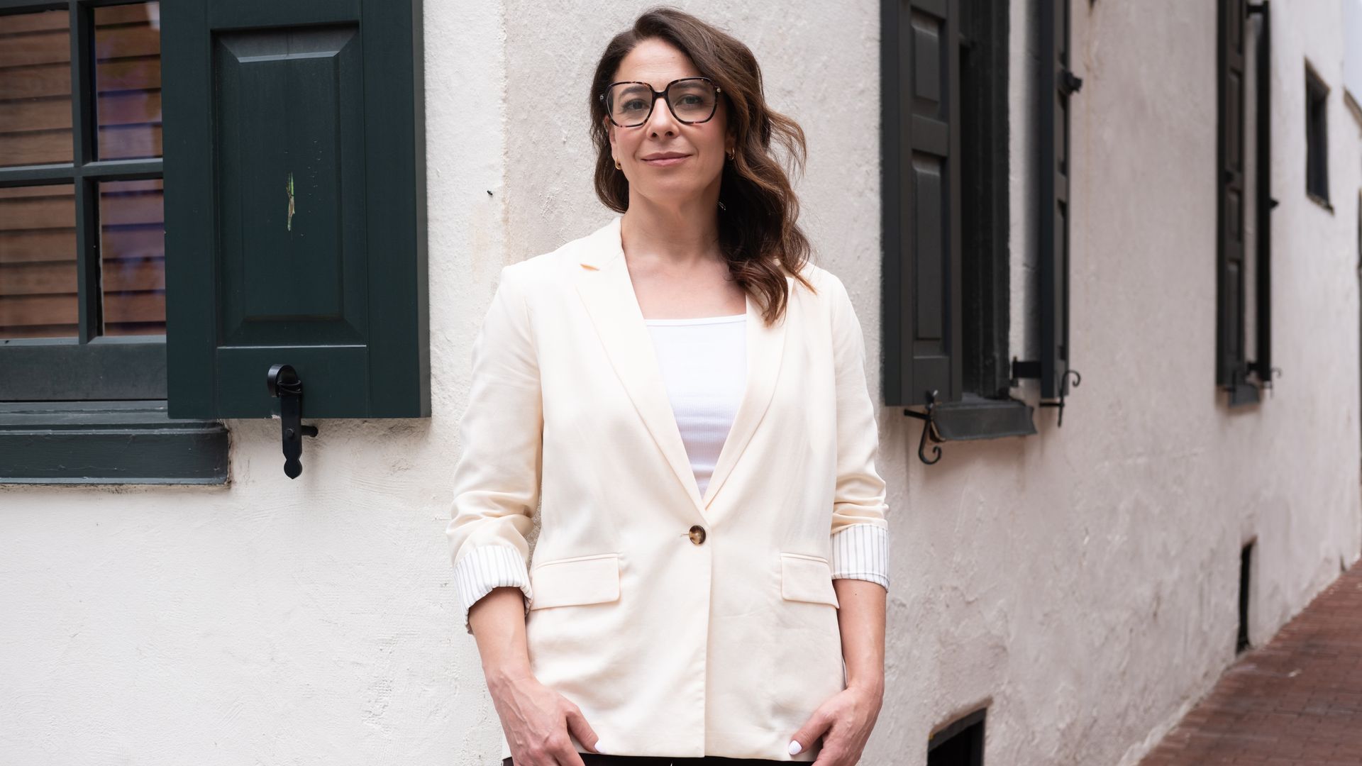 A woman with brown hair and large glasses stands outside near a white stucco wall with dark green shutters. She wears a cream blazer over a white top and dark pants, smiling slightly at the camera.