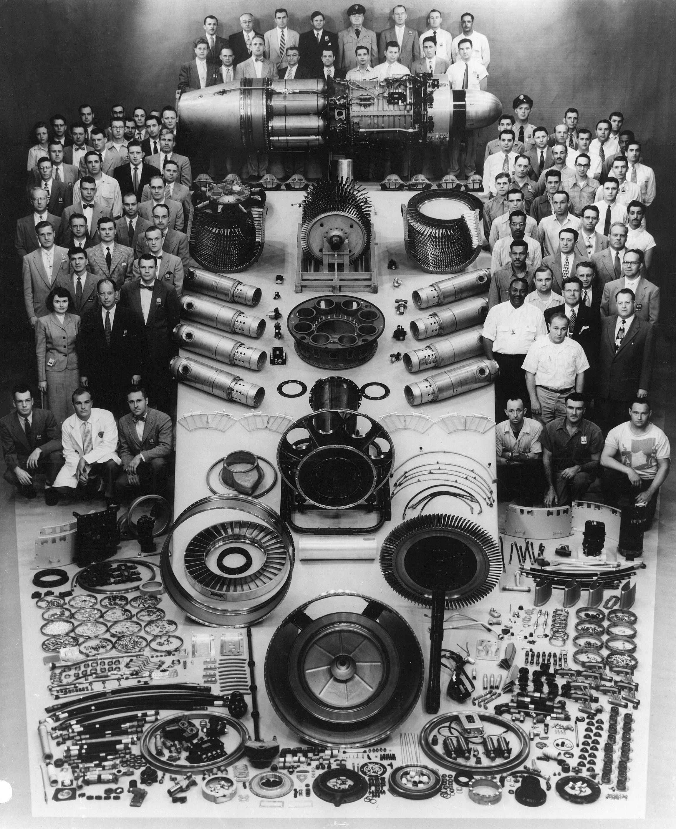 Black-and-white group photo of engineers and technicians surrounding a large rocket engine, with numerous disassembled components and circular parts laid out on tables.