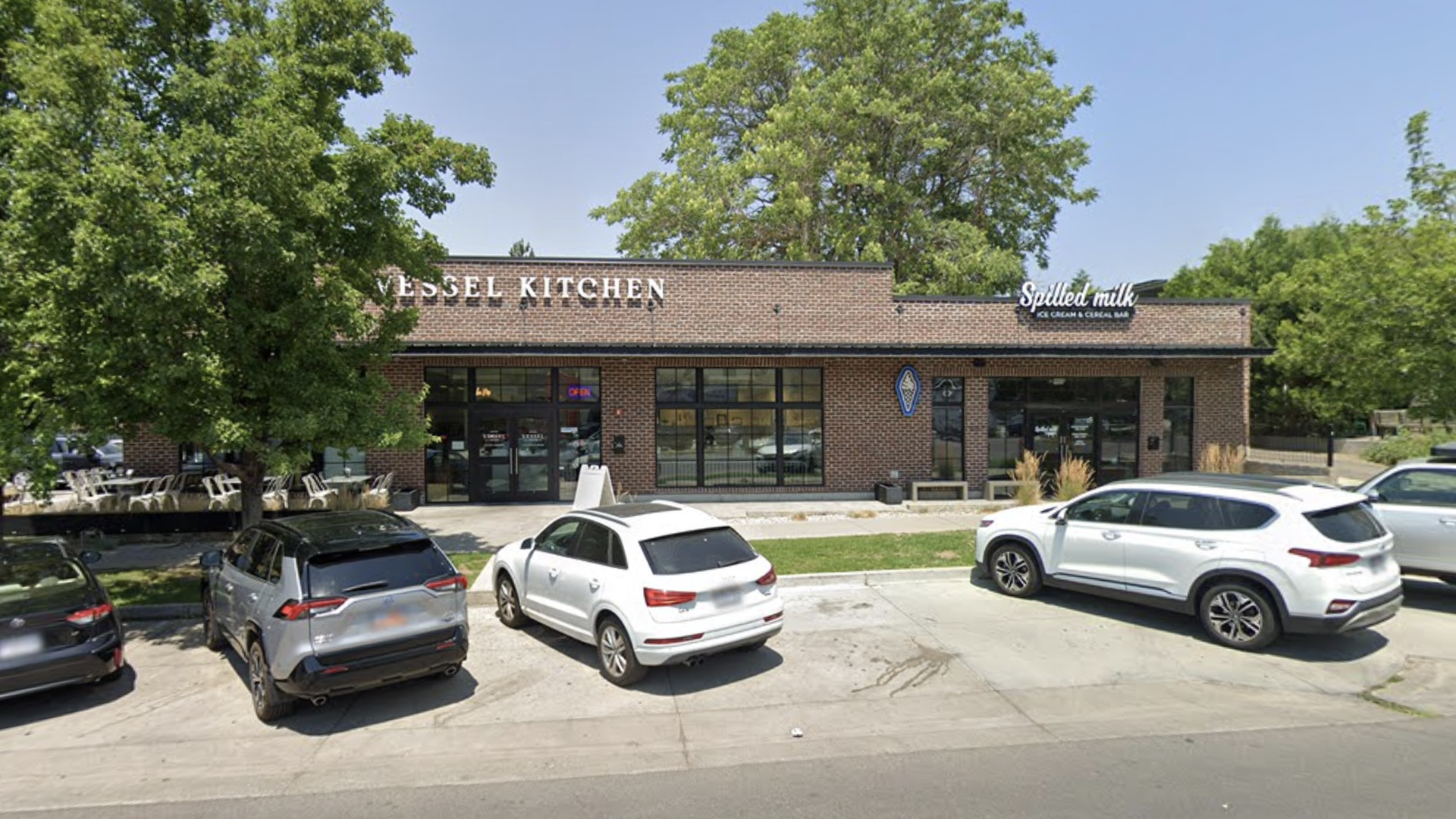 Brick storefront with two signs: Vessel Kitchen on the left and "Spilled Milk" on the right. Large windows, parked cars, leafy trees, and a bright blue sky.
