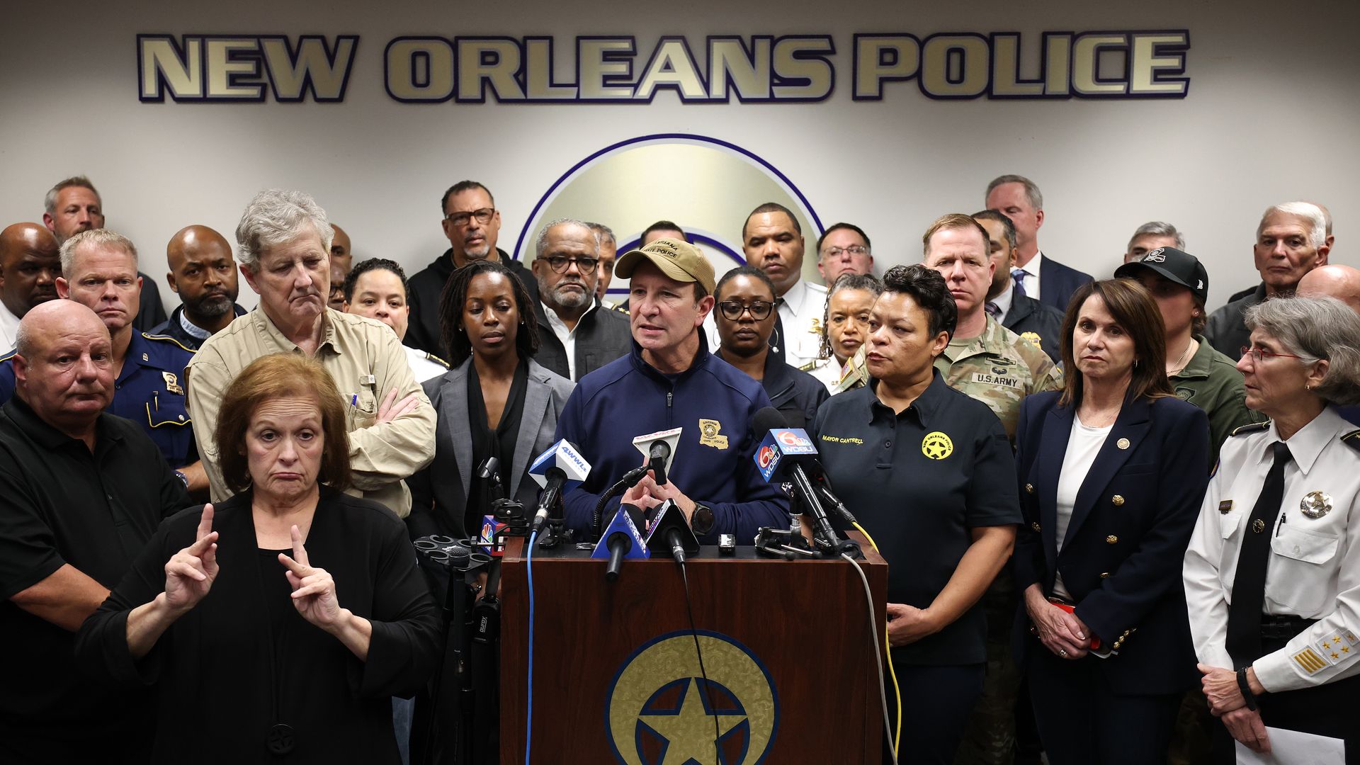 Gov. Jeff Landry speaks into a podium, flanked by dozens of officials, in the NOPD conference room.