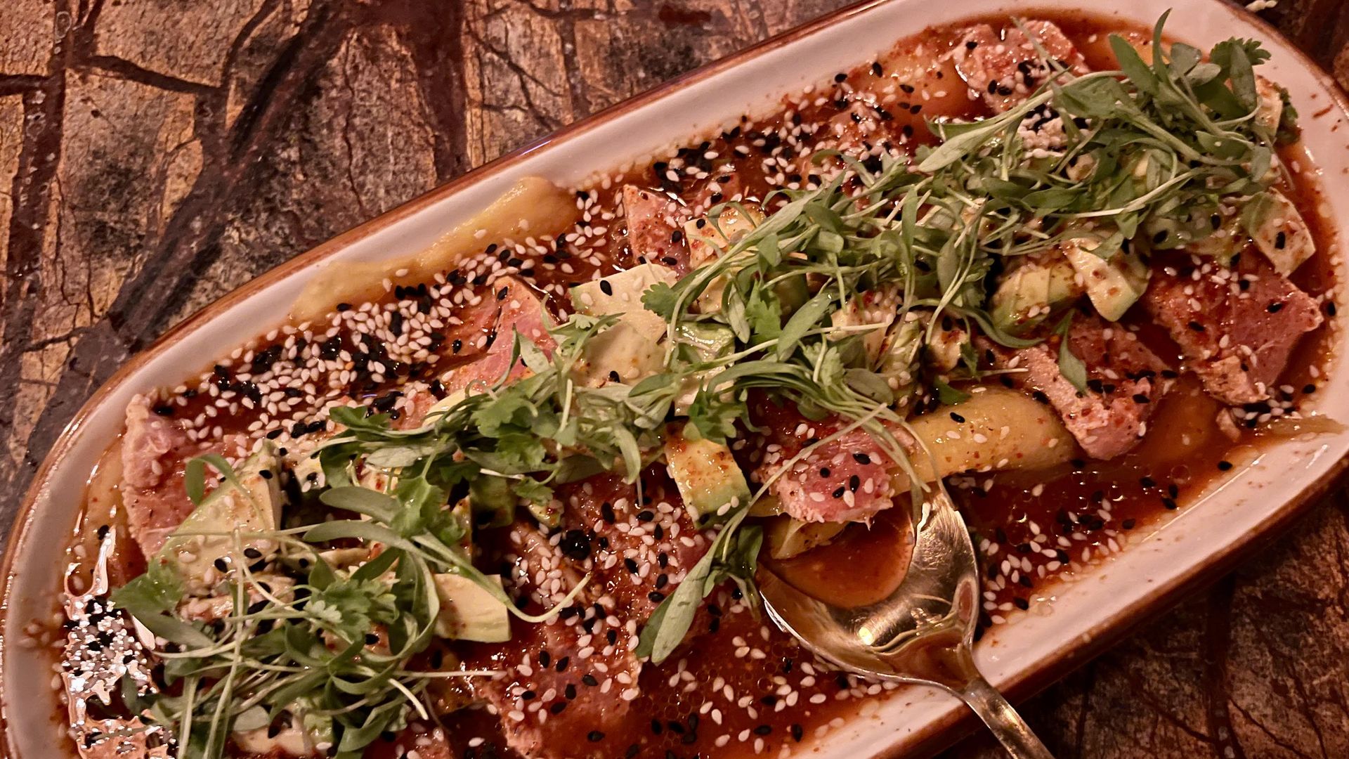 A long, oval plate is filled with red sauce, tuna and mango topped with greens and sesame seeds.