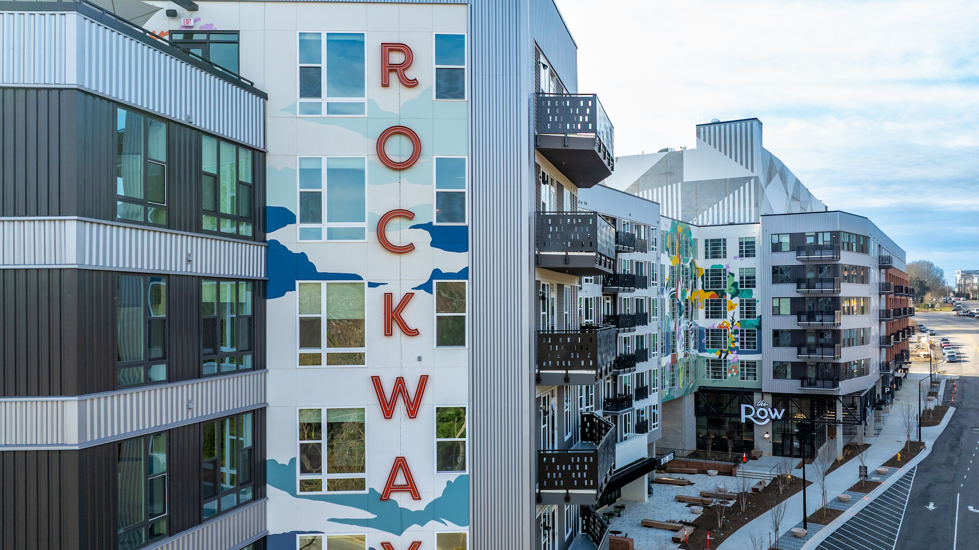 Modern apartment complex with large red vertical letters spelling "ROCKWAY" on white wall with blue cloud patterns. Balconies and a street with parked cars are visible.