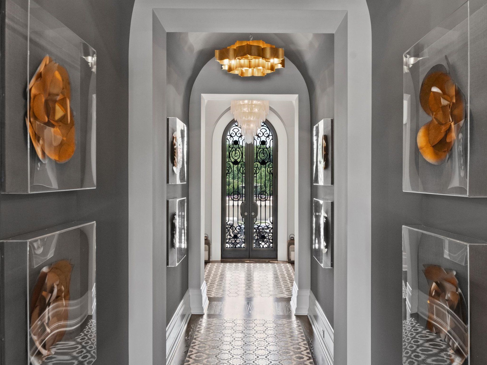 Long hallway with gray walls, decorative wall art in amber tones, patterned tile floor, two chandeliers—gold and white—and ornate black wrought iron double doors at the end.