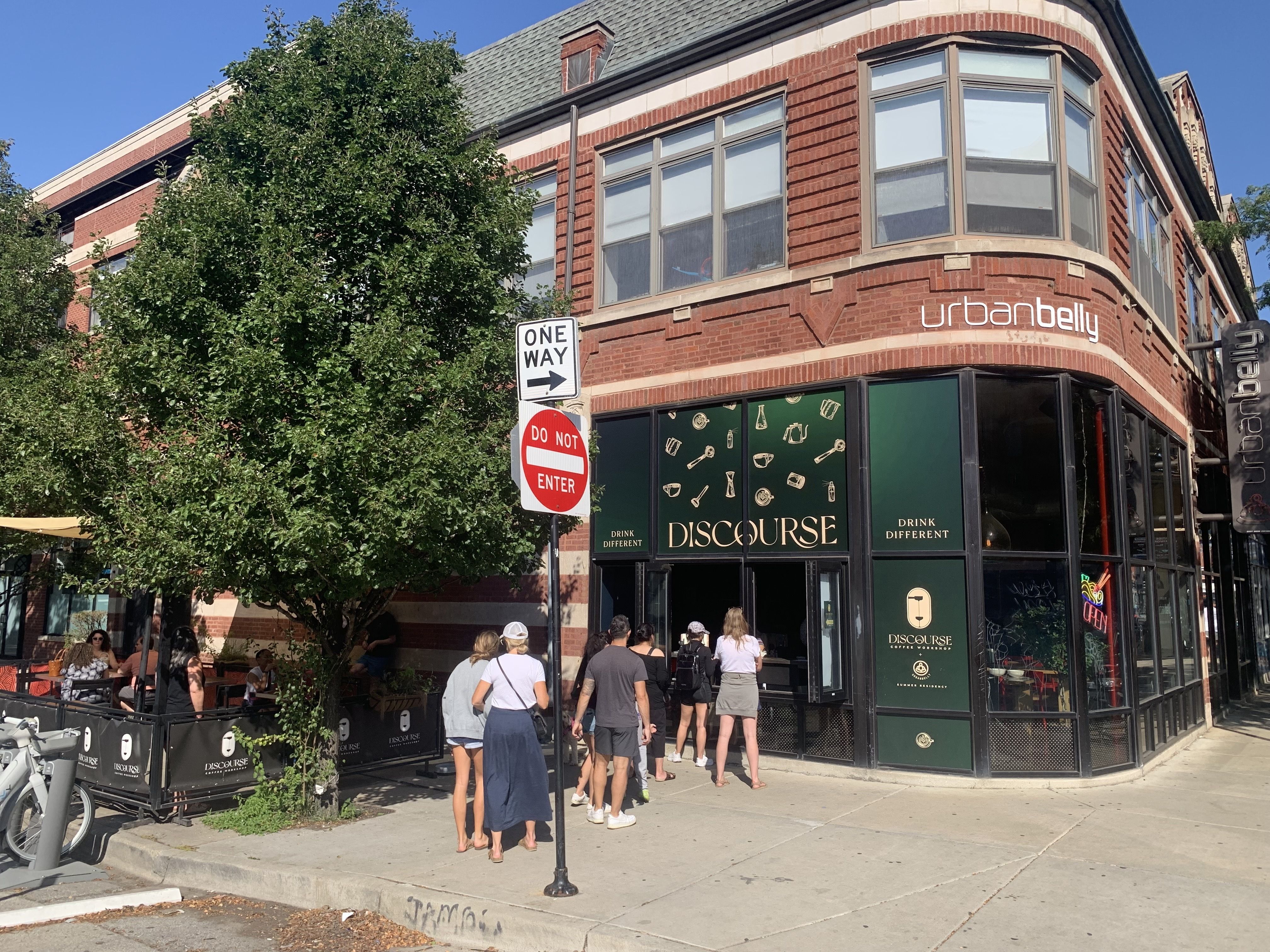 People standing in line outside Discourse Coffee Workshop, a corner café with green signage and city bikes parked nearby on a sunny day.