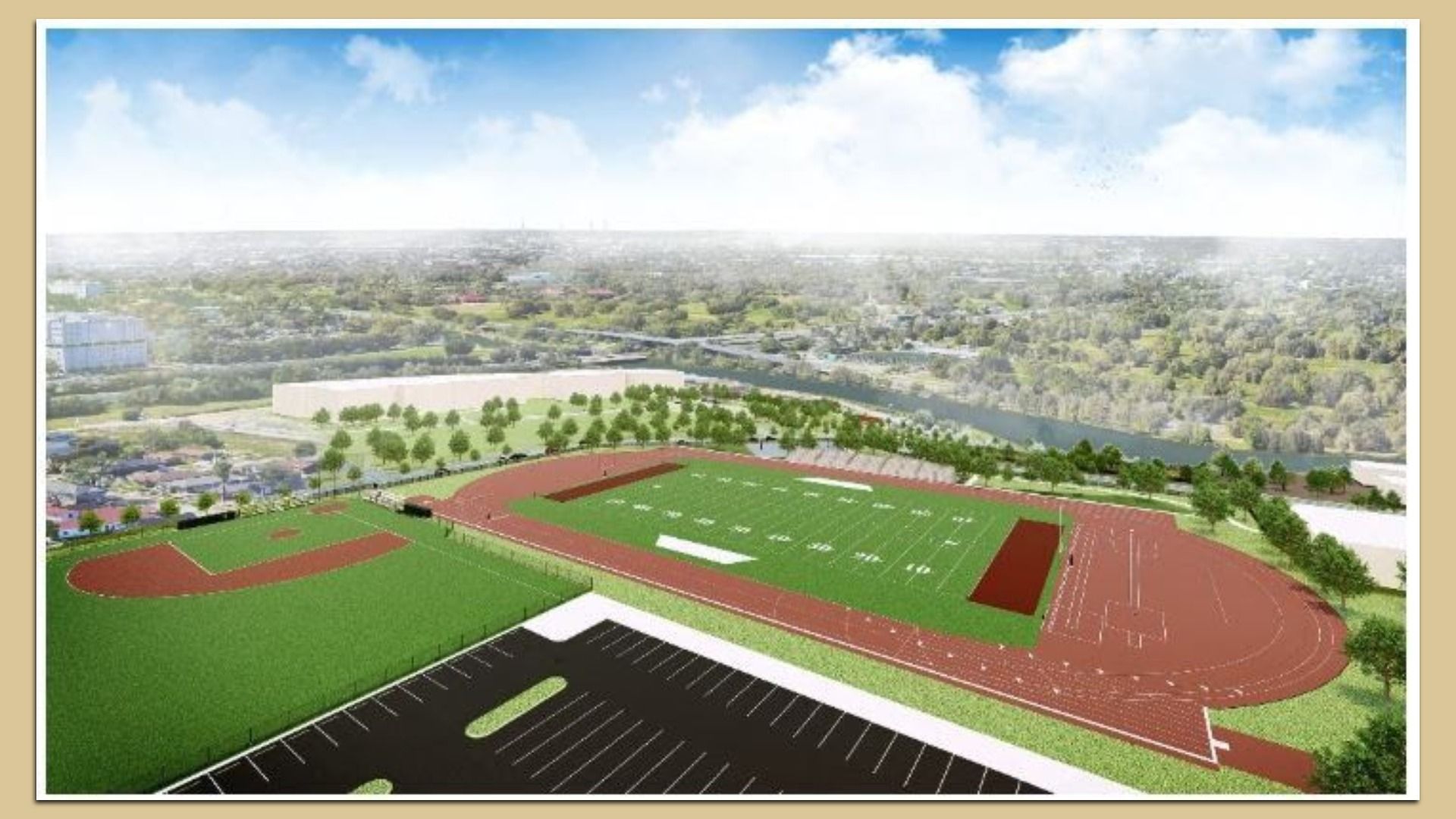 Aerial view of a sports complex with a green football field, red running track, adjacent baseball field, parking lot, trees, and a river in the background under a partly cloudy sky.