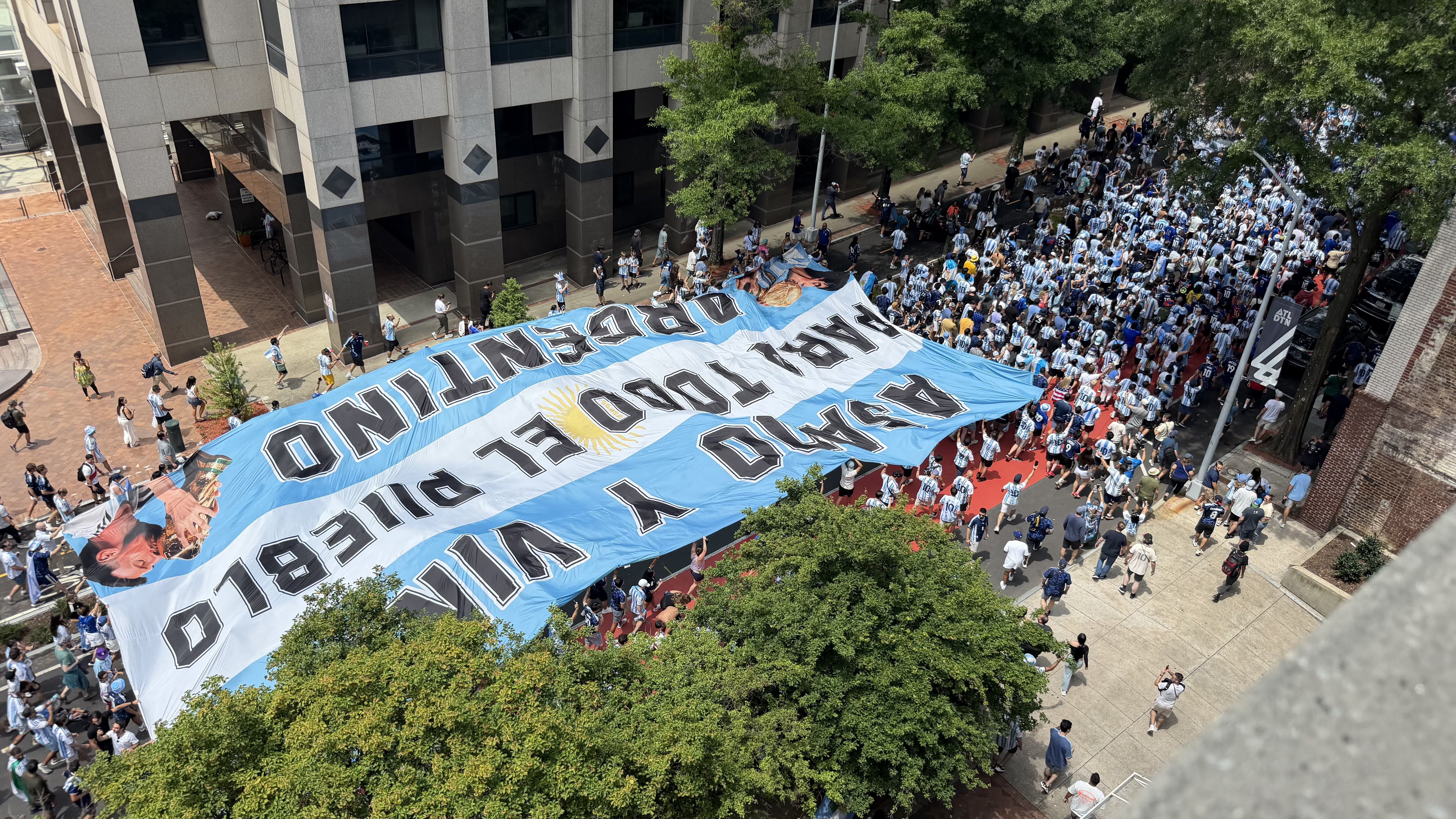 An aerial view of a city street parade showing a large blue-and-white banner with a sun emblem; crowds in blue-and-white striped outfits fill the road between modern buildings and trees.