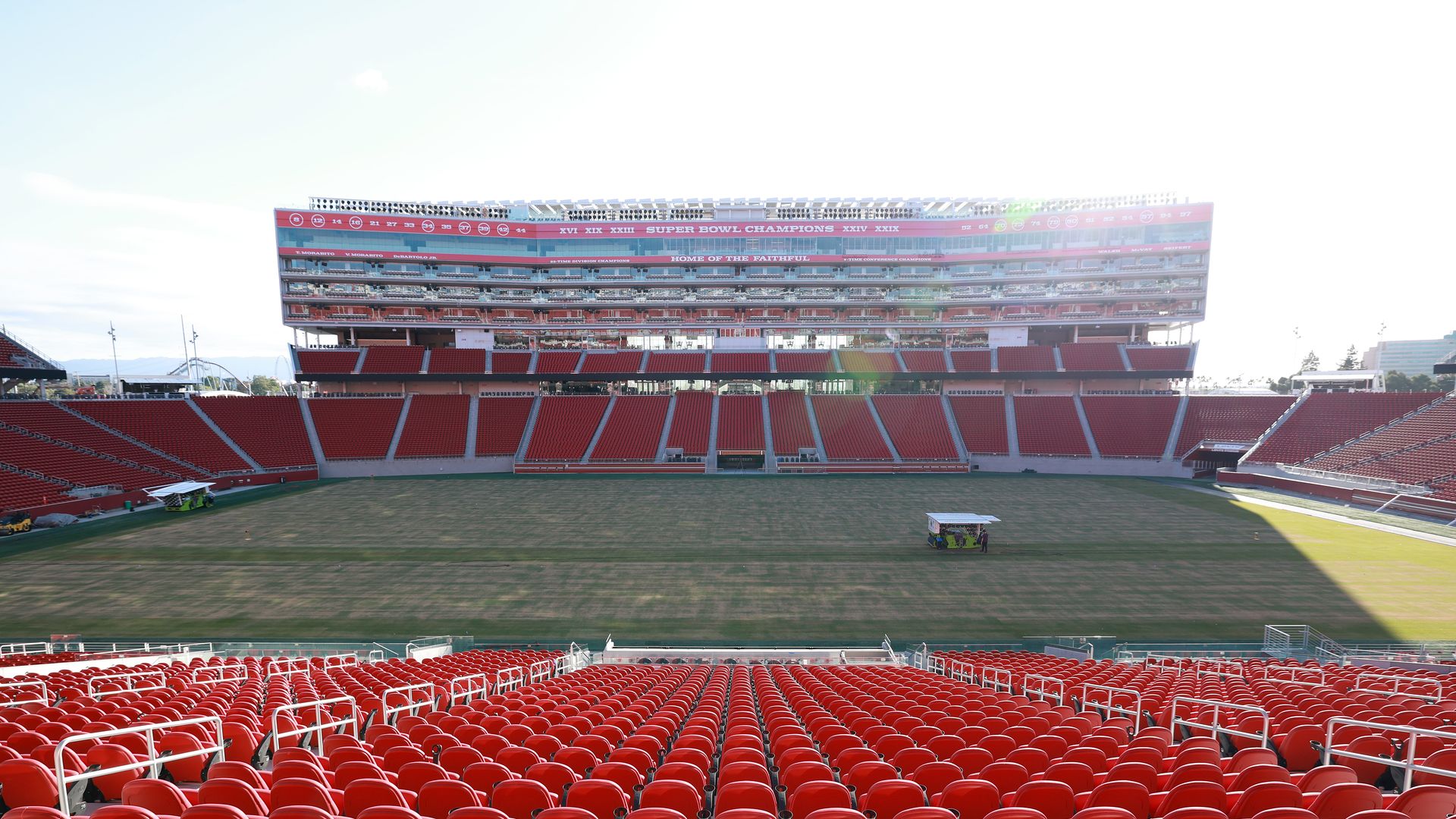 Wide view of a large football stadium with bright red seats and a multi-level press box tower across the field. A banner reads "SUPER BOWL CHAMPIONS"; empty stands and maintenance carts on the grass.