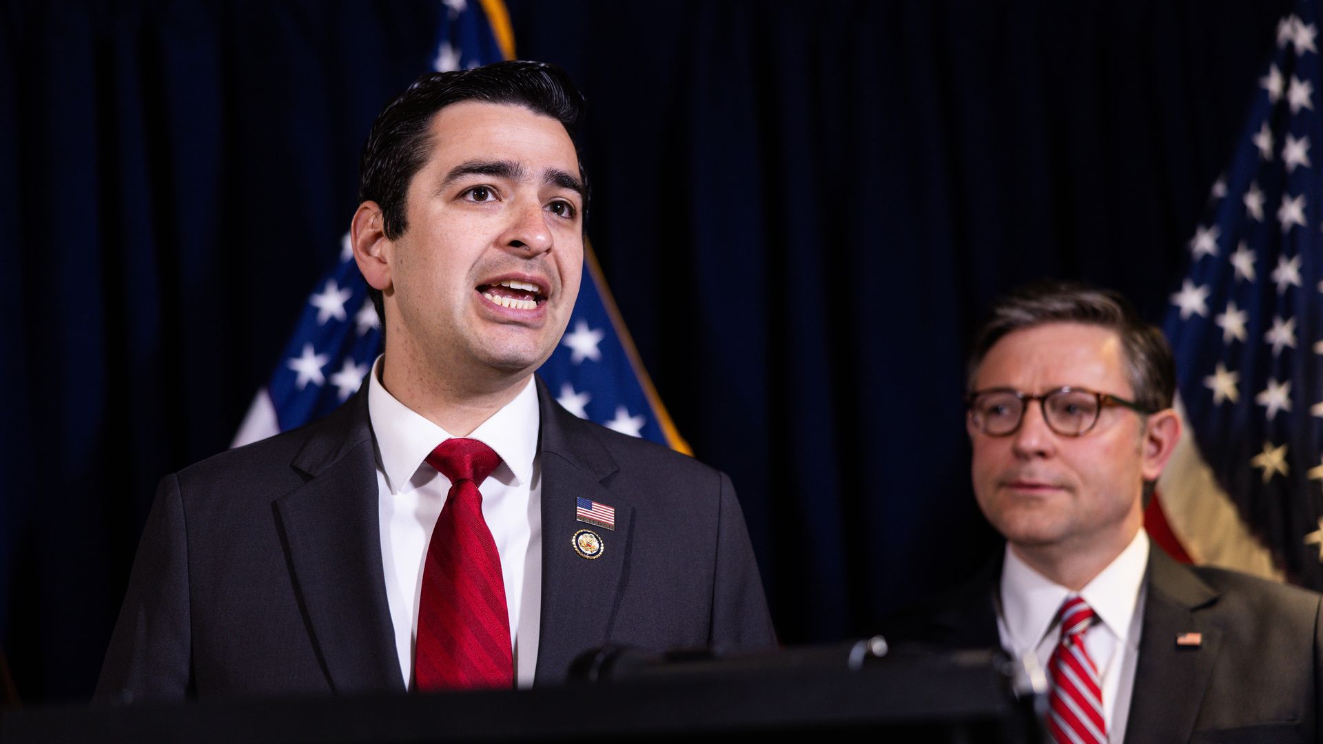 A man in a suit with a necktie speaks while on a lectern. 