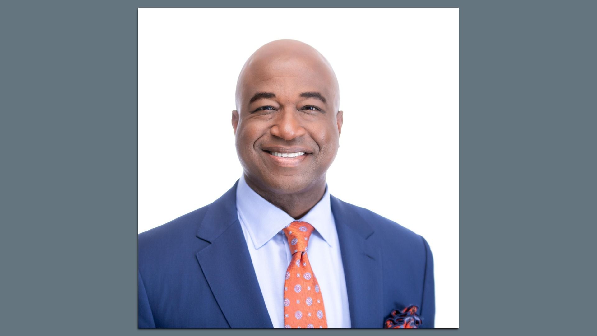 Smiling bald man in a navy suit, light blue shirt, and orange patterned tie with a pocket square, posed against a white background.