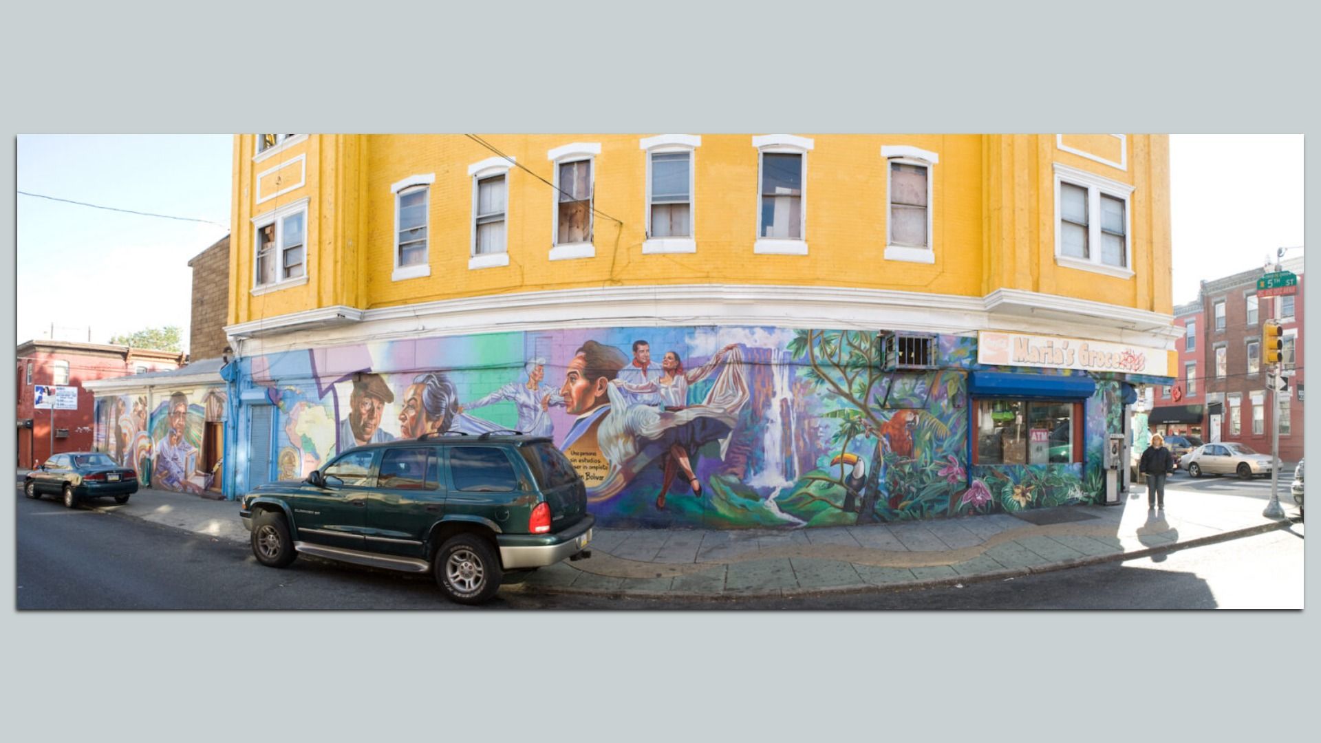 Mural-covered yellow corner building with people and tropical scenes. A blue storefront reads "Mardi's Grocery." A green SUV and a car are parked along the curb as a pedestrian walks by.