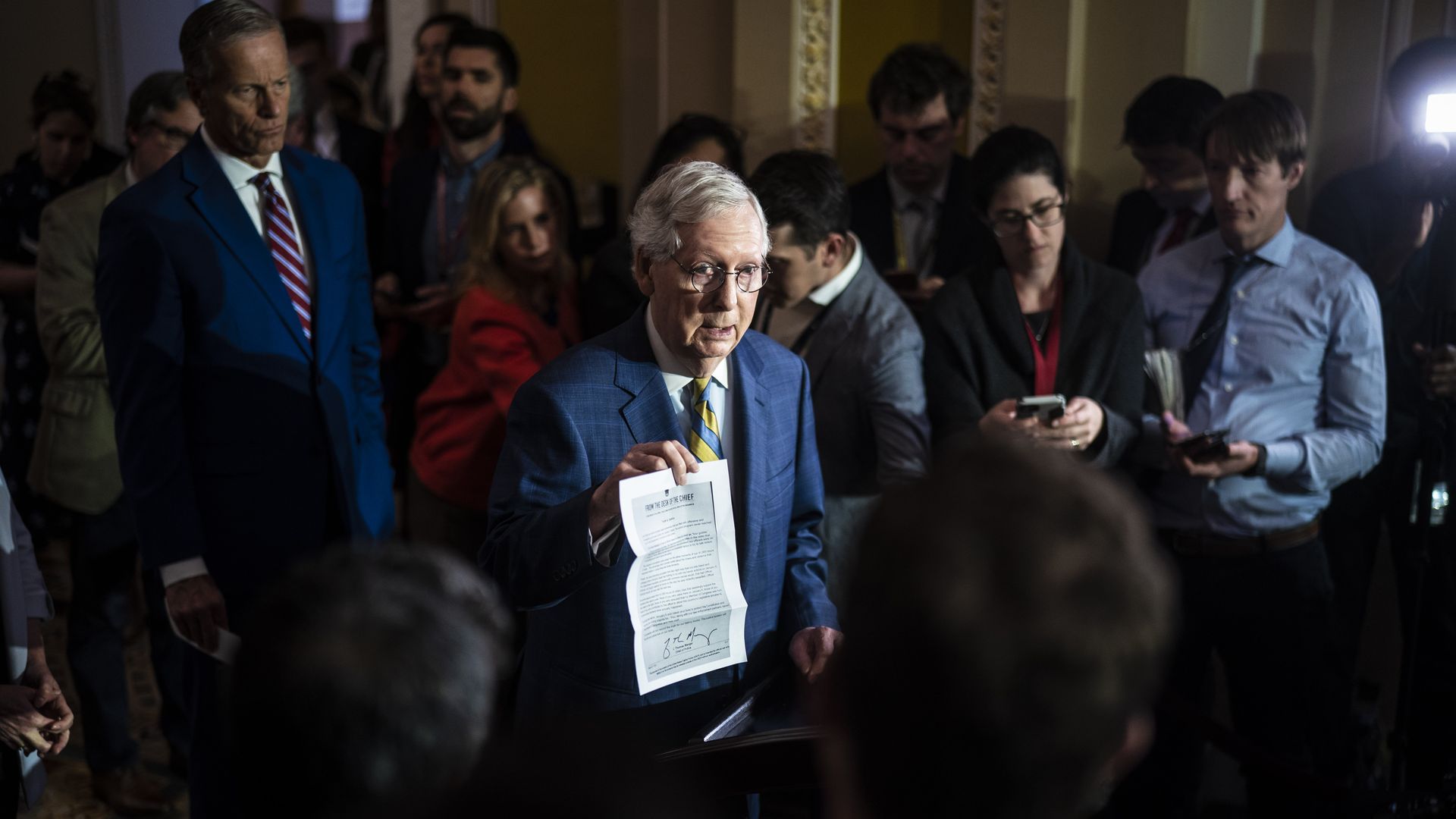 Sen. Mitch McConnell, wearing a blue suit and white shirt, holds up a piece of paper while surrounded by colleagues and Capitol Hill reporters.