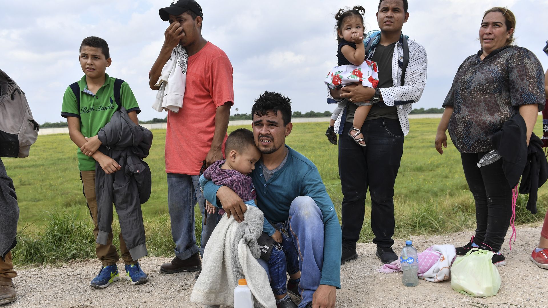In this image, a line of migrants stand outside, several holding small children.