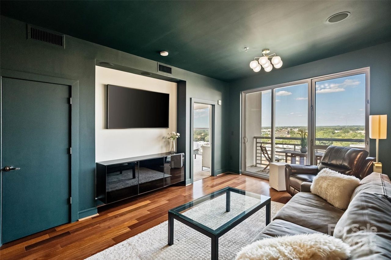Modern living room with dark teal walls and ceiling, large window with balcony view, leather couch, glass coffee table, wooden floor, and wall-mounted TV above black cabinet.