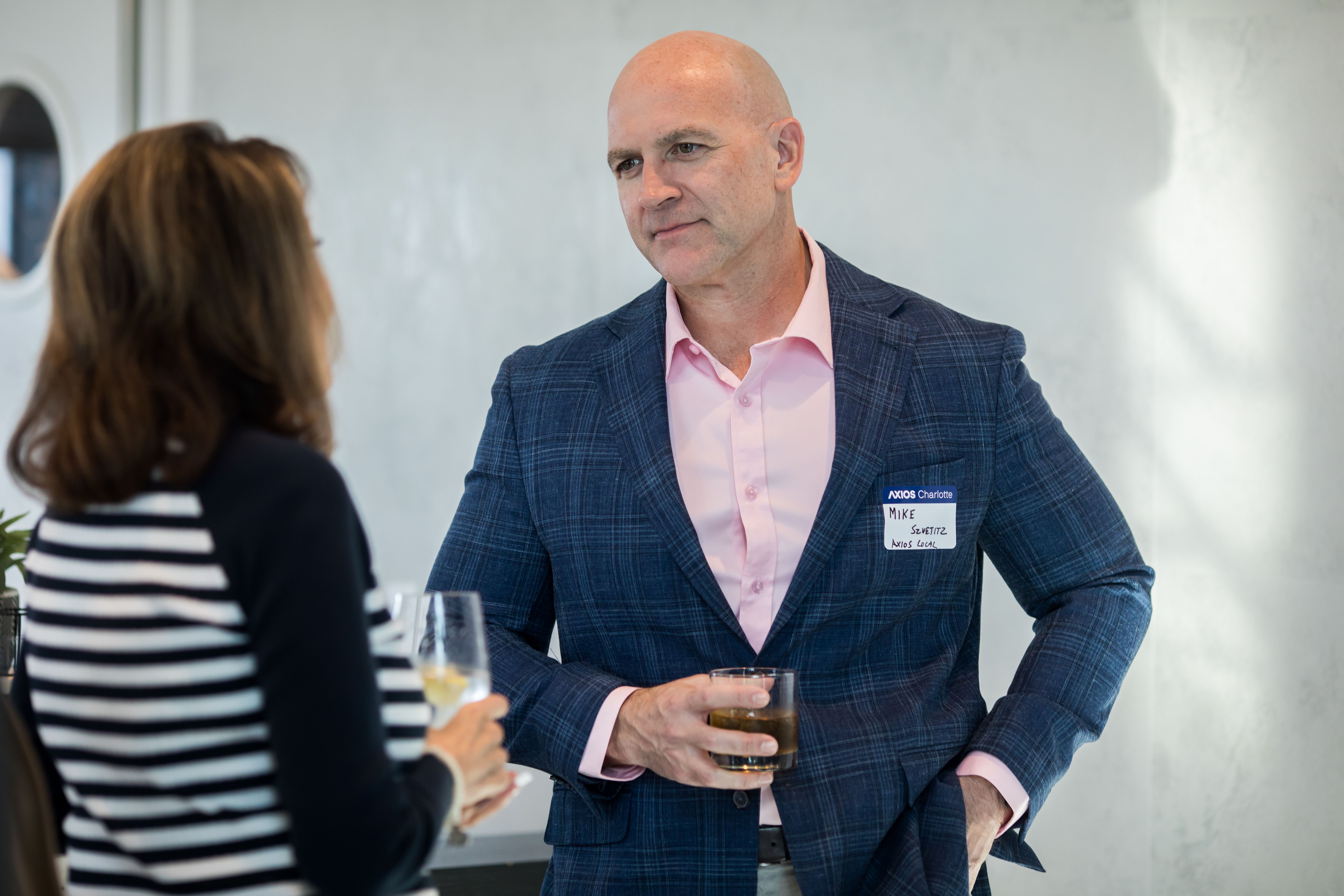 Bald man in a pink shirt and blue plaid blazer chats with a person in a striped top; he wears a name badge and holds a glass of dark beverage in a bright indoor setting.