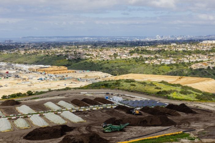 A birdseye view of a landfill and compost facility with several rows of piles of waste, and a city in the distance.