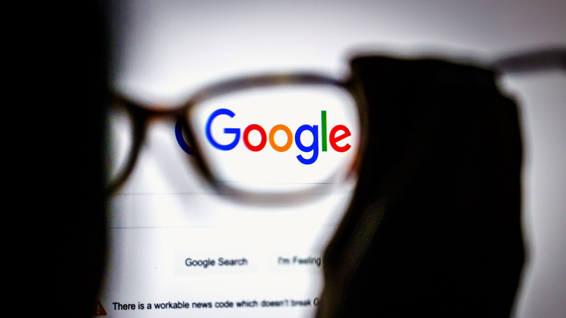 Photo of a screen showing the Google search page as a person holds up glasses that magnify the Google logo