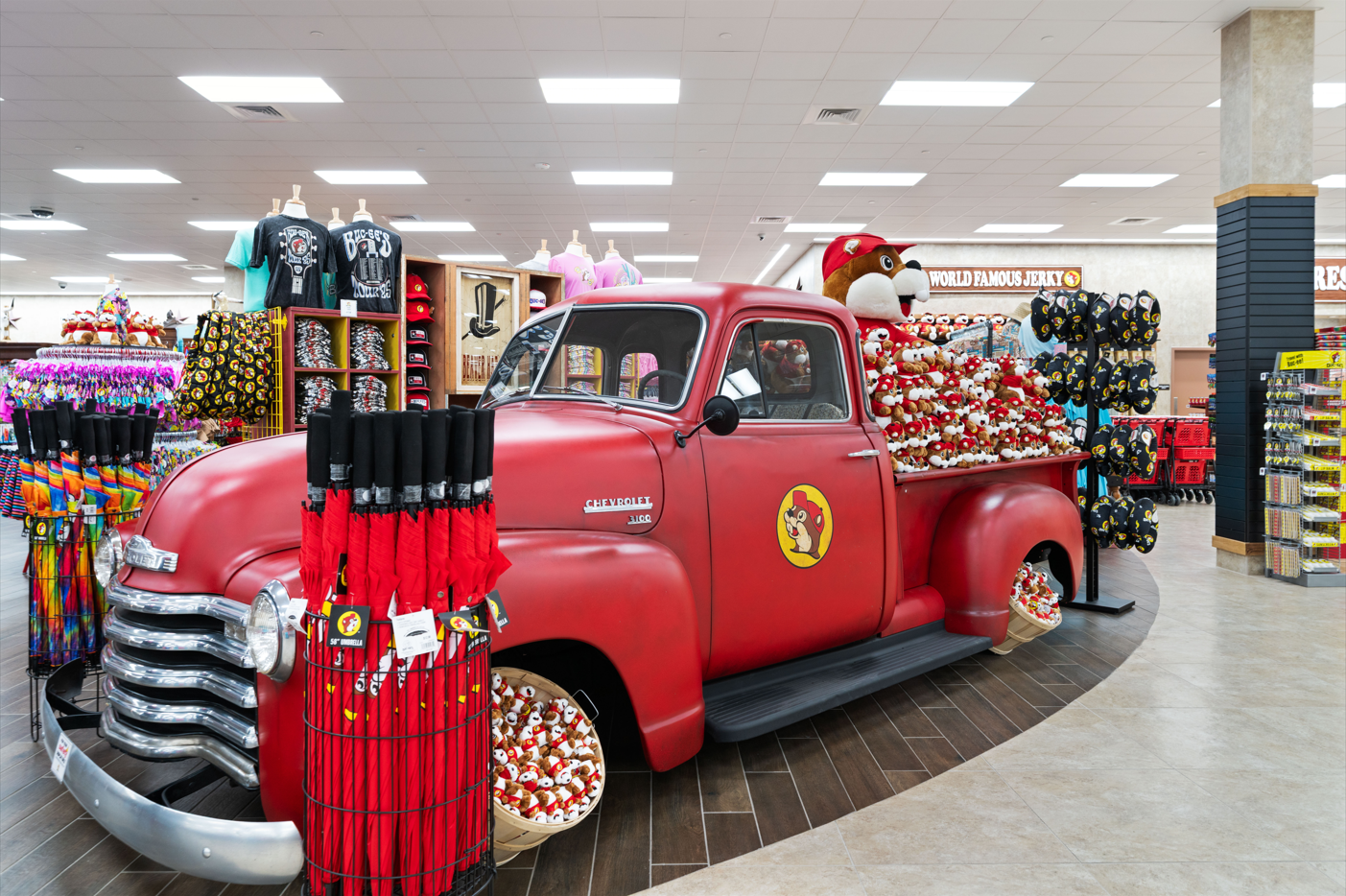 Red vintage Chevrolet pickup inside the Buc-ee's store in Huber Heights, surrounded by clothing racks, umbrellas, hats, and shelves of plush bears and souvenirs on the truck bed.