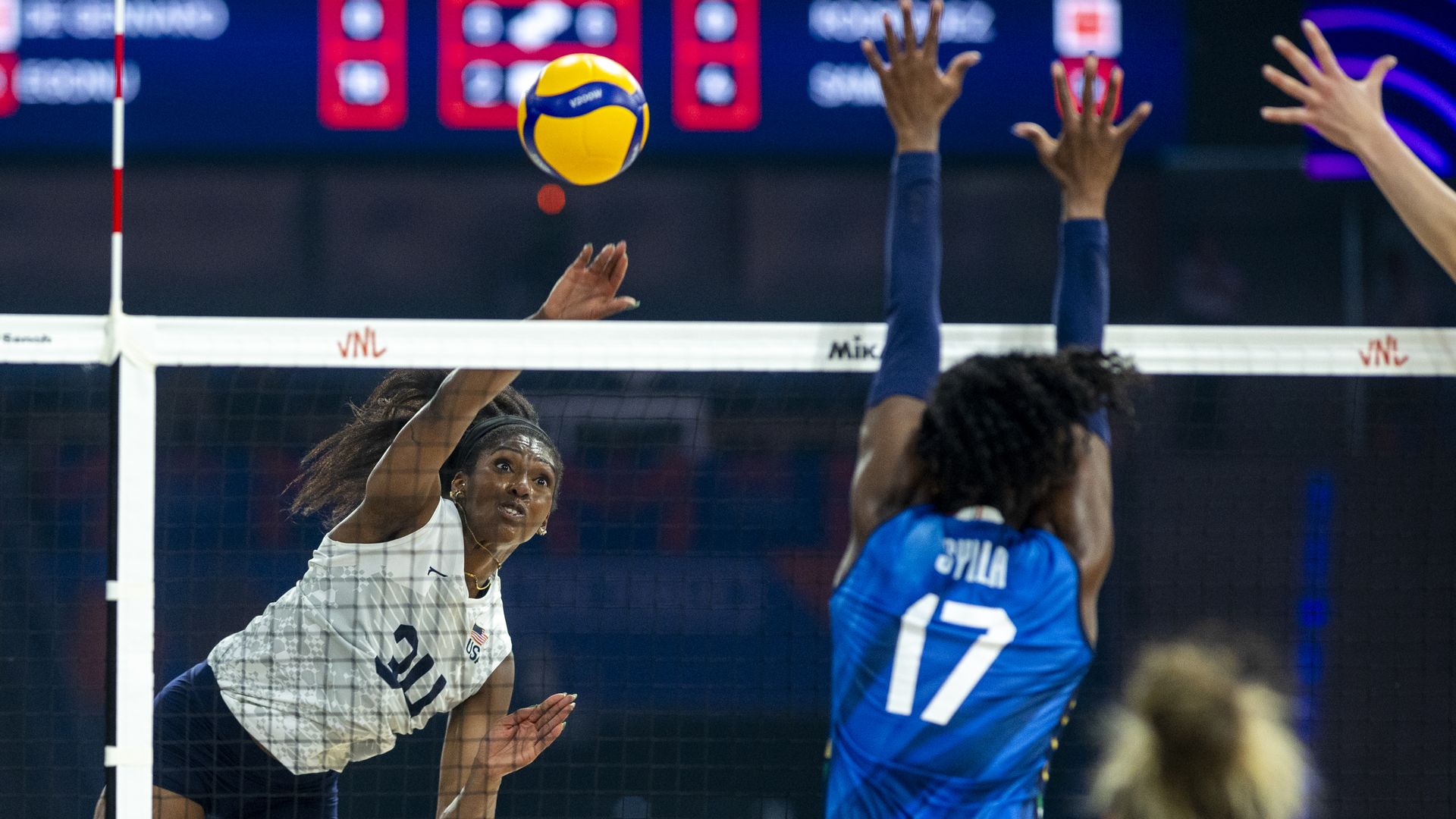 Female volleyball player in white jersey #21 spikes ball over net against opponent in blue jersey #17 blocking during a match with scoreboard blurred in the background.