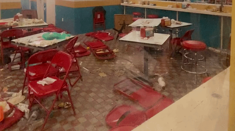 a video from outside a window of the inside of a restaurant. red chairs and food are strewn on the floor, Tables are moved out of whack