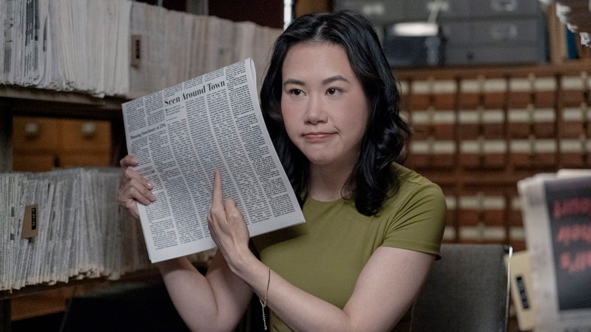 Woman with black hair in a green shirt sitting in a room full of archives, pointing at a newspaper article titled "Seen Around Town" with a neutral expression.