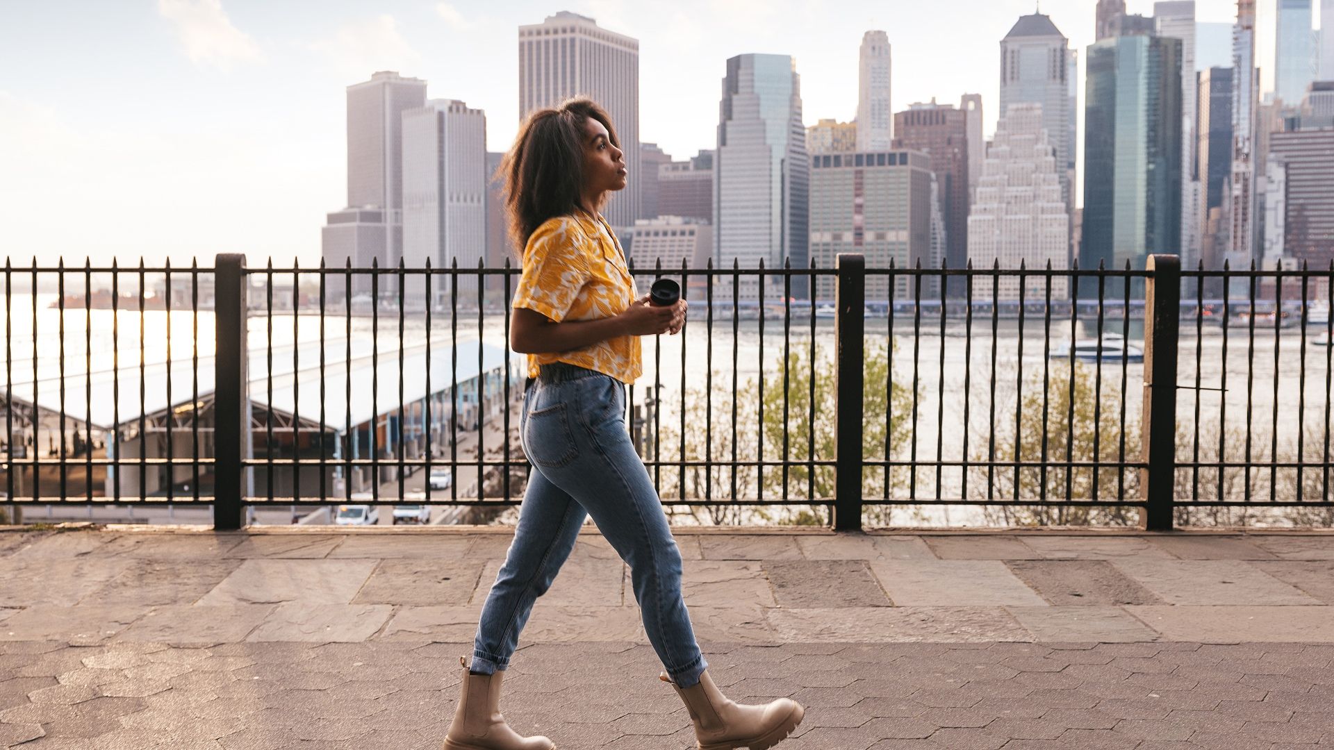 Woman in yellow shirt and jeans walking by railings with a city skyline and river in the background during daylight, holding a cup in her hands.