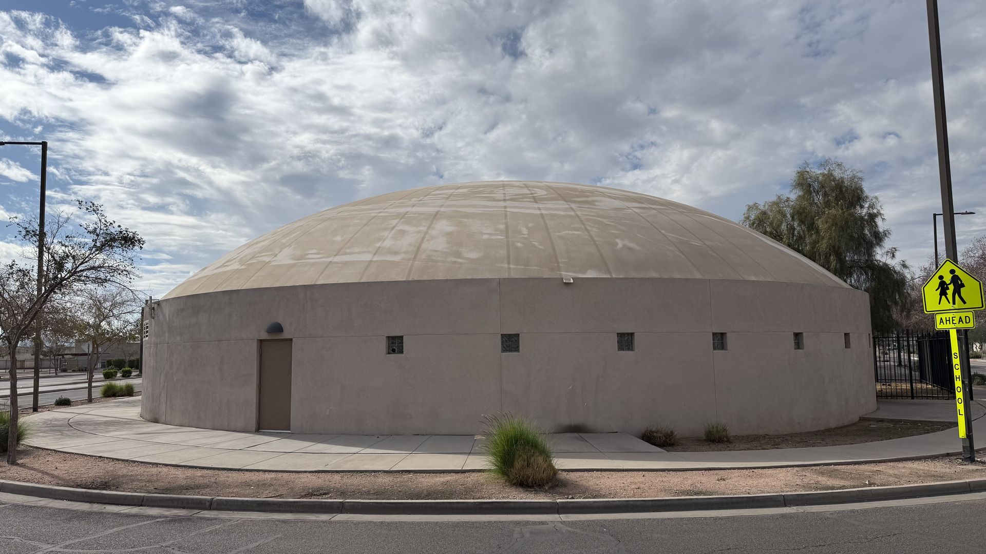 A large beige domed building with a smooth curved roof under a partly cloudy blue sky, small square windows near the base, a single door, shrubbery, and a bright yellow "School Ahead" pedestrian sign nearby.