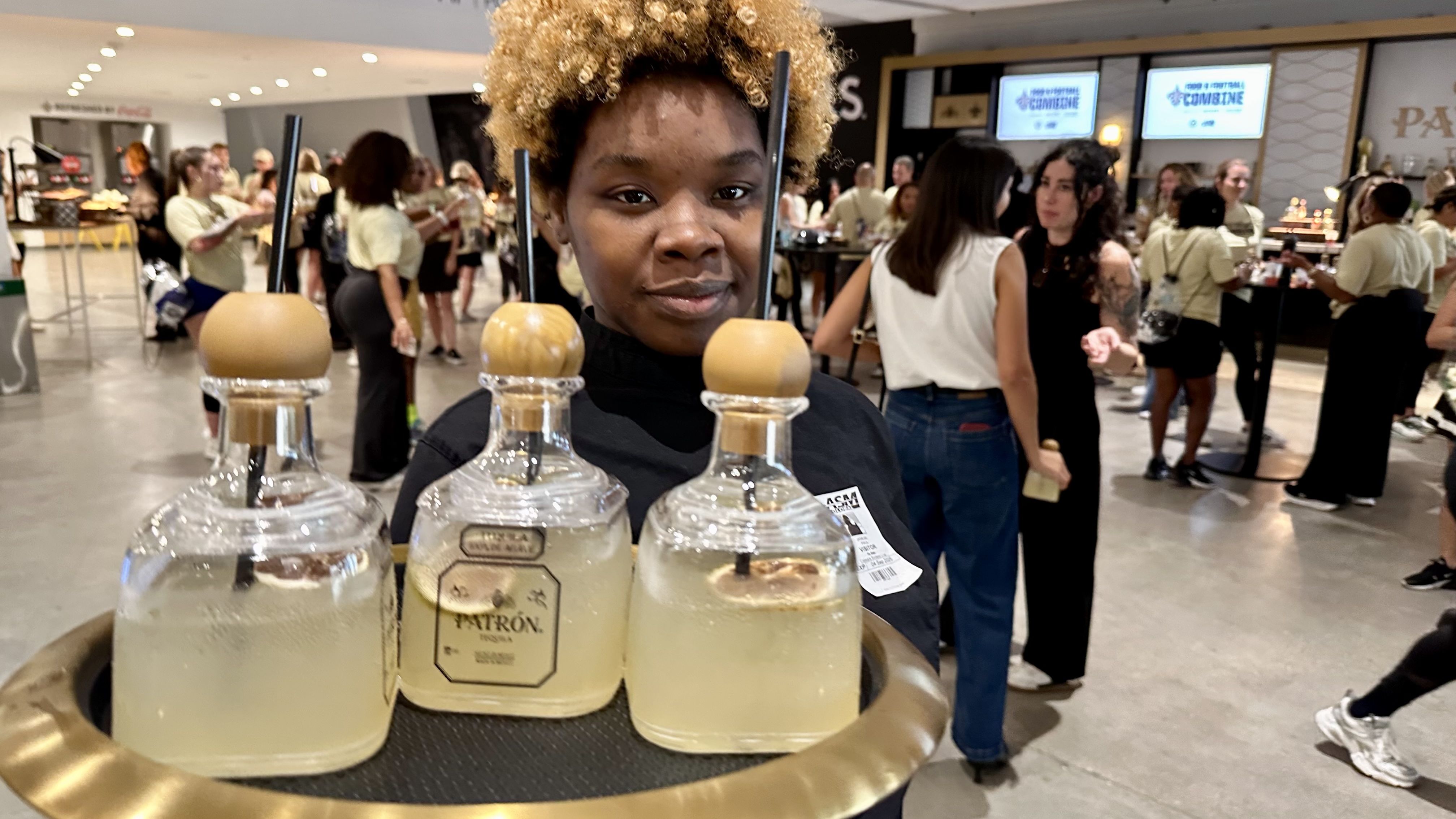 Young person with curly hair carrying a tray with three Patron tequila cocktails in clear bottles with round wooden tops at a busy indoor event with people socializing in the background.