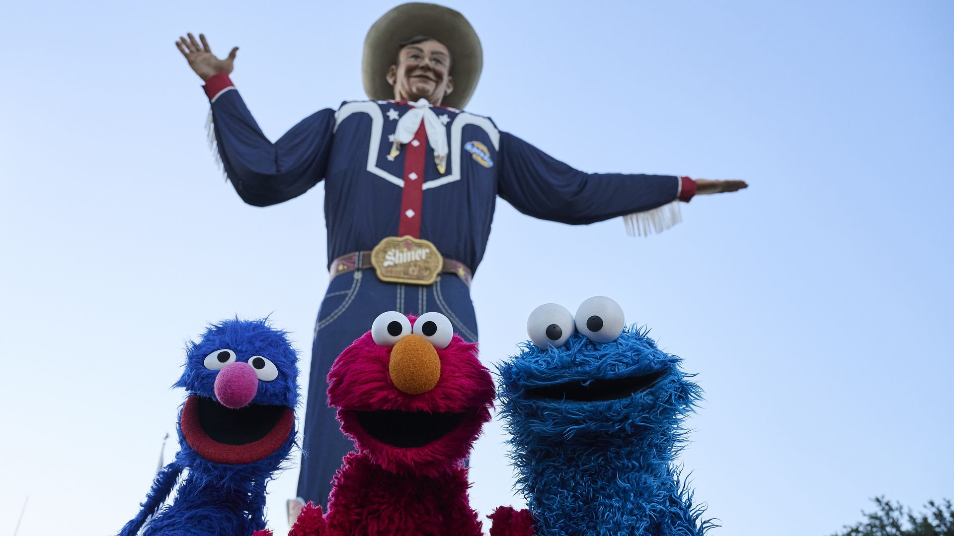 Three Sesame Street characters, Grover, Elmo, and Cookie Monster, pose smiling with a giant cowboy statue in the background against a clear blue sky.