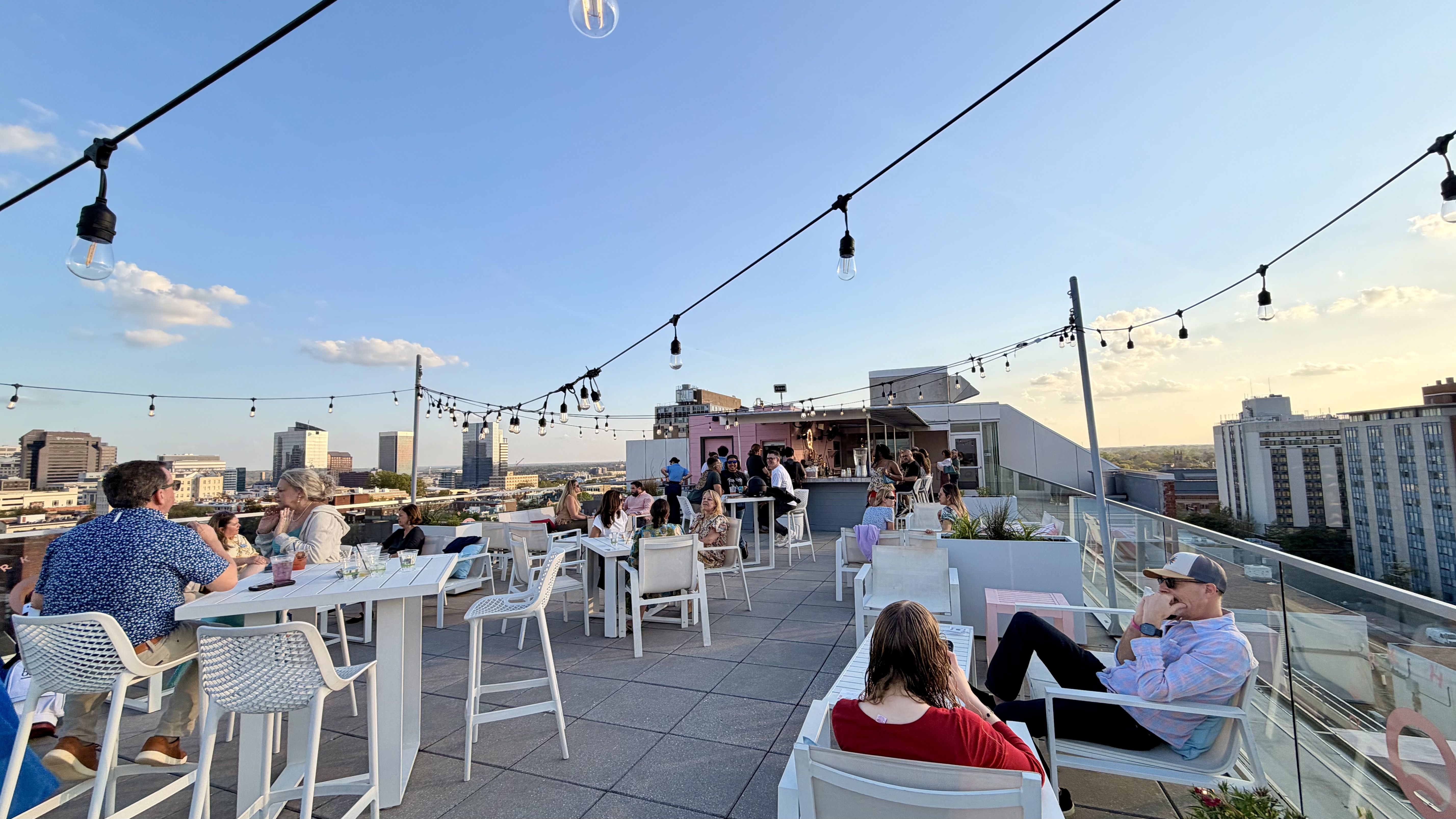 Rooftop dining terrace with string lights, white tables and chairs, glass railing, and groups of people enjoying drinks as a city skyline glows at sunset.