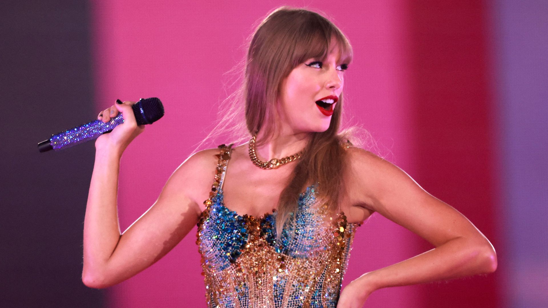 Taylor Swift smiles sassily while looking over her left shoulder and holding a bejeweled microphone with her right hand