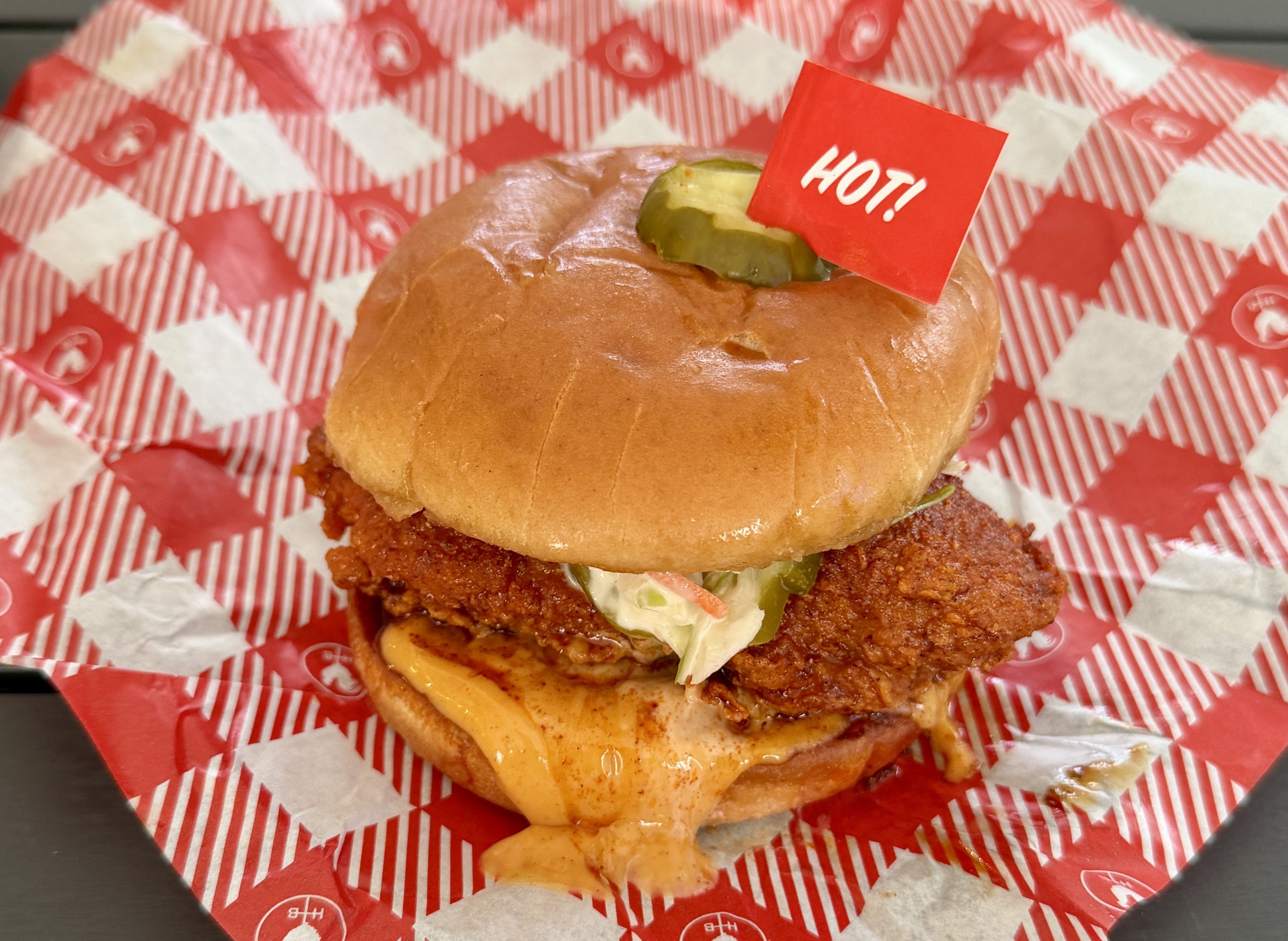 Crispy fried chicken sandwich with melted cheese, pickle on top, orange sauce dripping, served on red-and-white checkered paper with a red HOT! flag.
