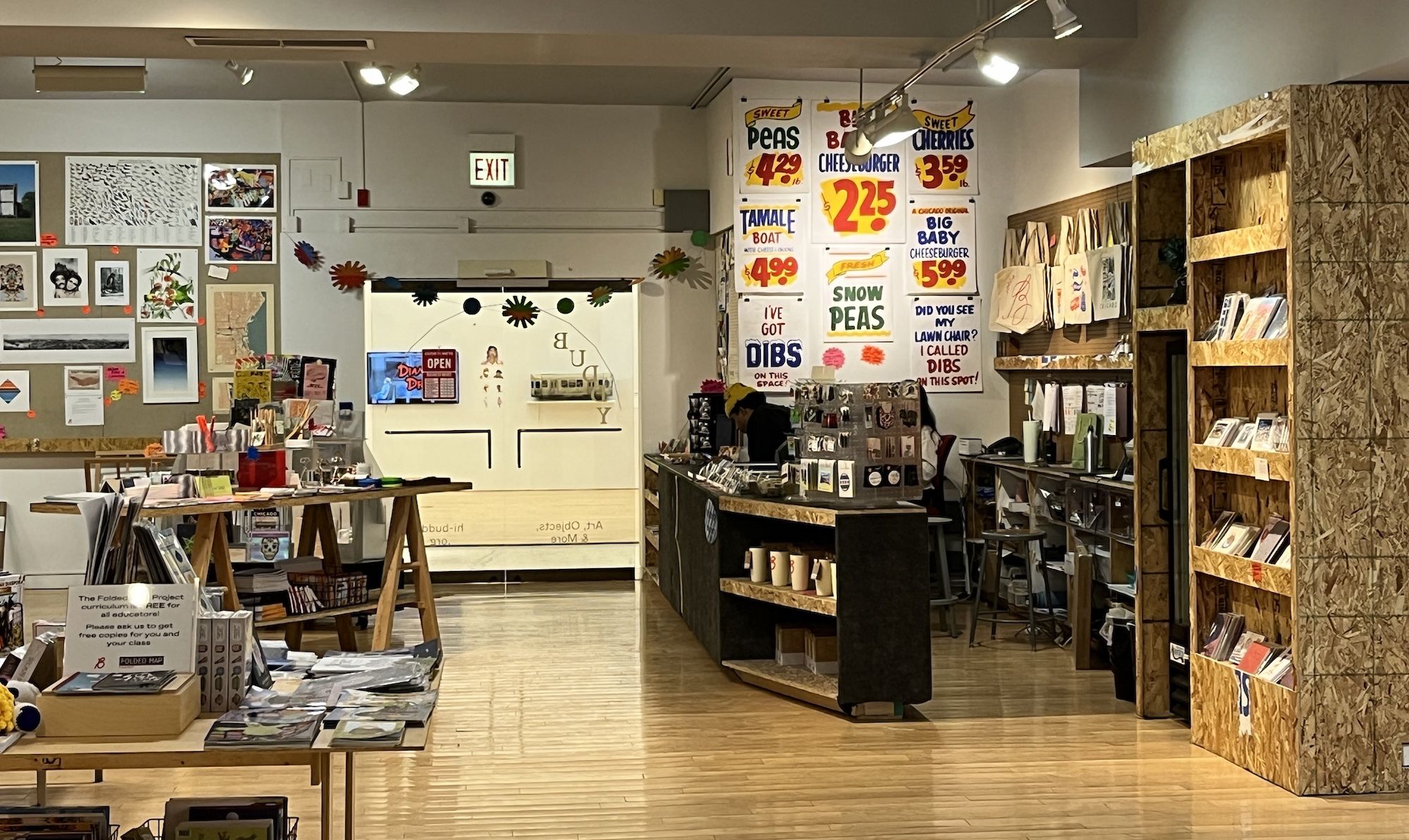 Interior of a well-lit art shop with wooden floors, colorful sale signs, art prints on walls, shelves holding books and merchandise, and two people behind counters.