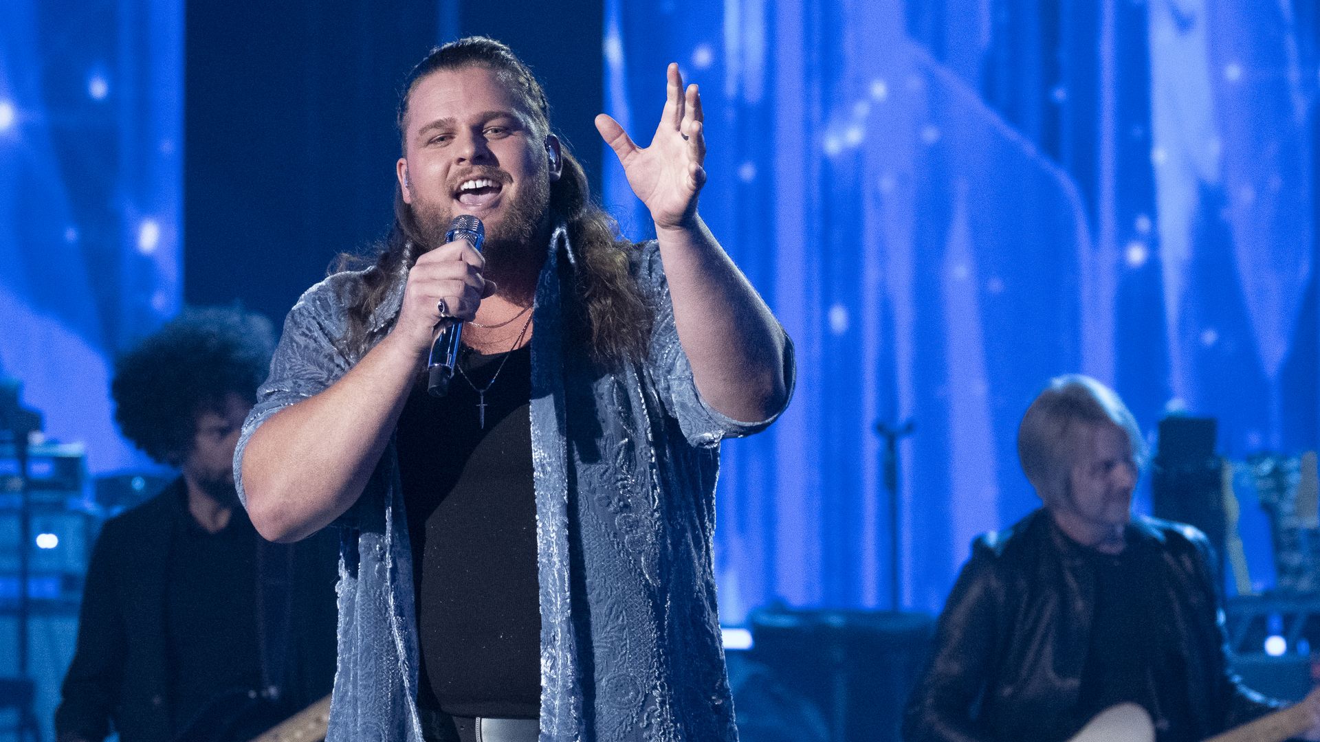 Male singer on stage singing into a microphone with one hand raised; wearing a black shirt under a blue patterned open shirt. Blue stage lighting with band members visible in the background.
