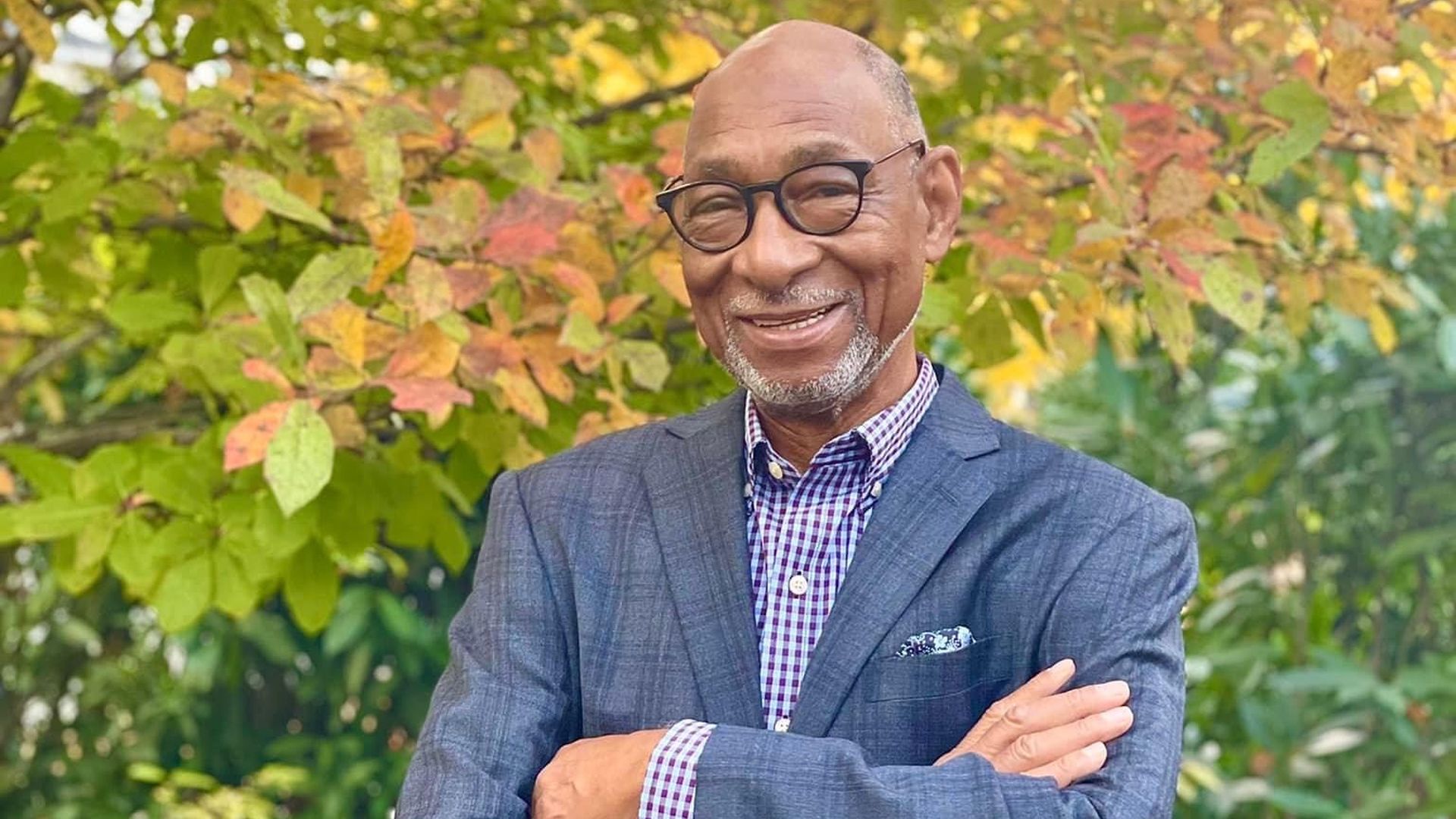 Smiling older man with glasses wearing a blue-gray blazer and checkered shirt, arms crossed, standing in a garden with autumn foliage—yellow, orange, and green leaves behind him.