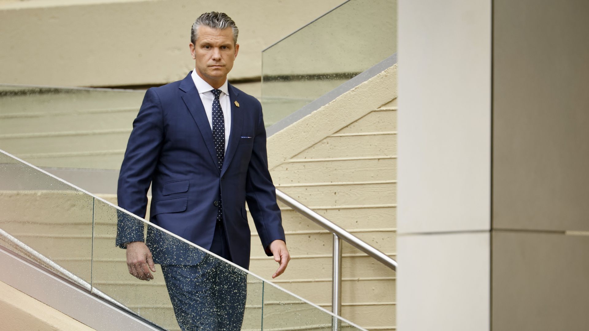 Man in a navy suit and polka-dot tie stands on a glass-railed staircase inside a beige building, looking at the camera.