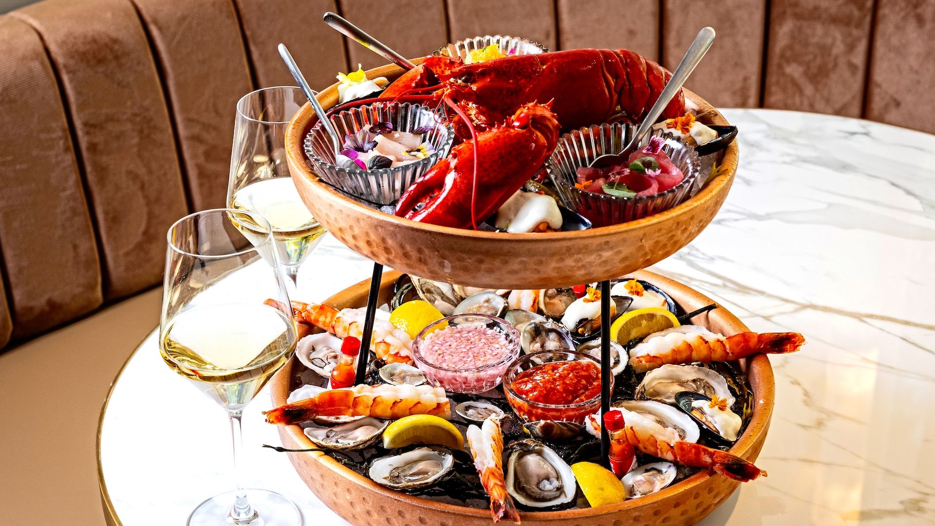 Two-tiered seafood platter with a red lobster, shrimp, oysters, and sauces on a white marble table, accompanied by two glasses of white wine.