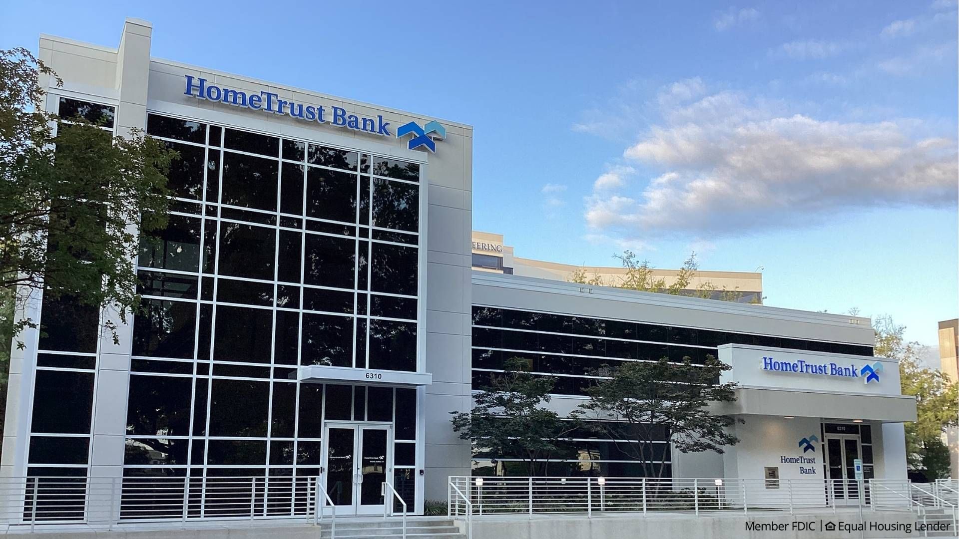 Sunlit modern bank building with large black-tinted glass panels, white framework, and blue "HomeTrust Bank" signs; trees in front, a wheelchair ramp, and a clear blue sky.