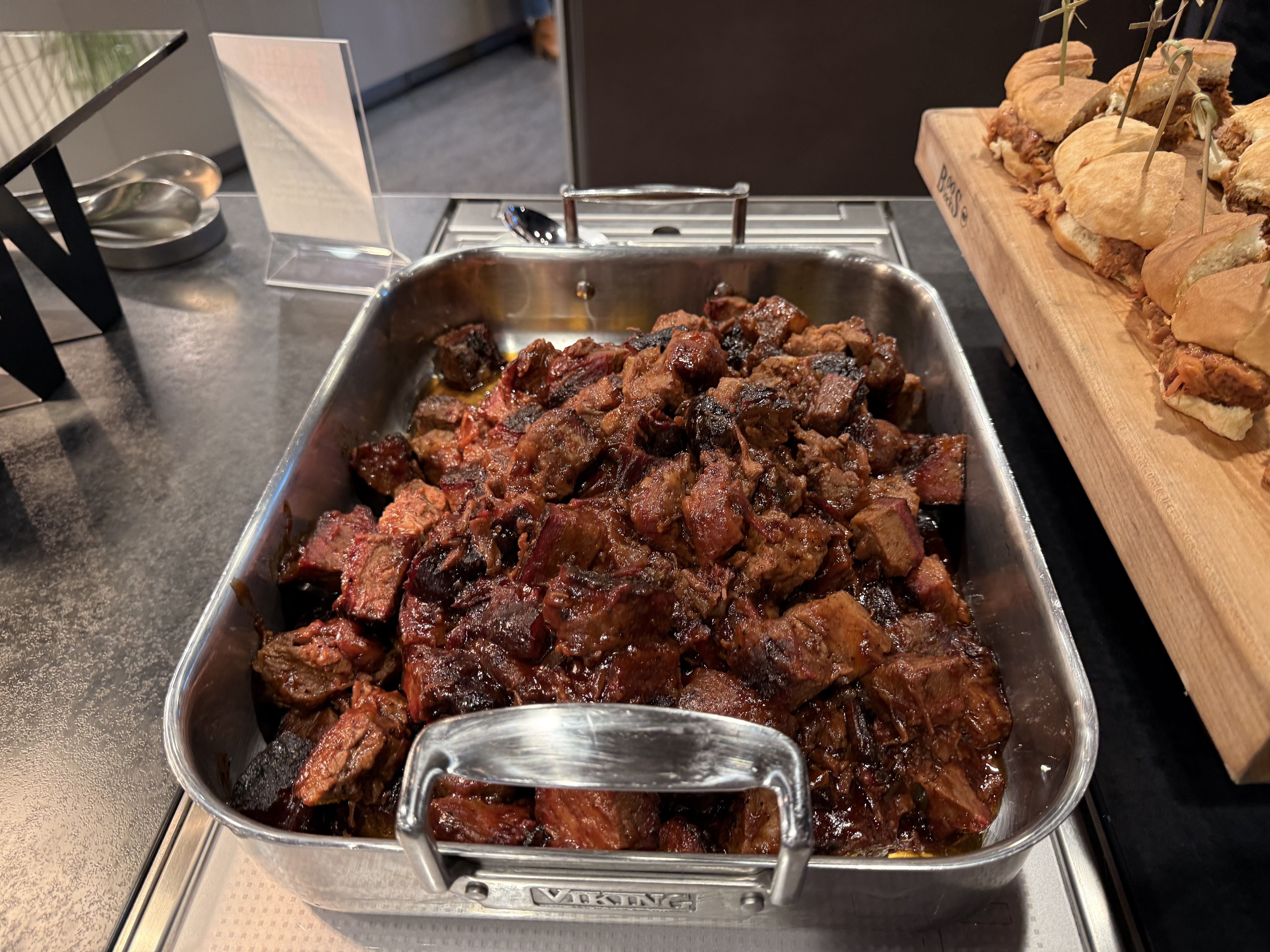 Buffet scene with a metal tray of braised, caramelized beef chunks in juices; to the right, a wooden board piled with small sandwiches on skewers.