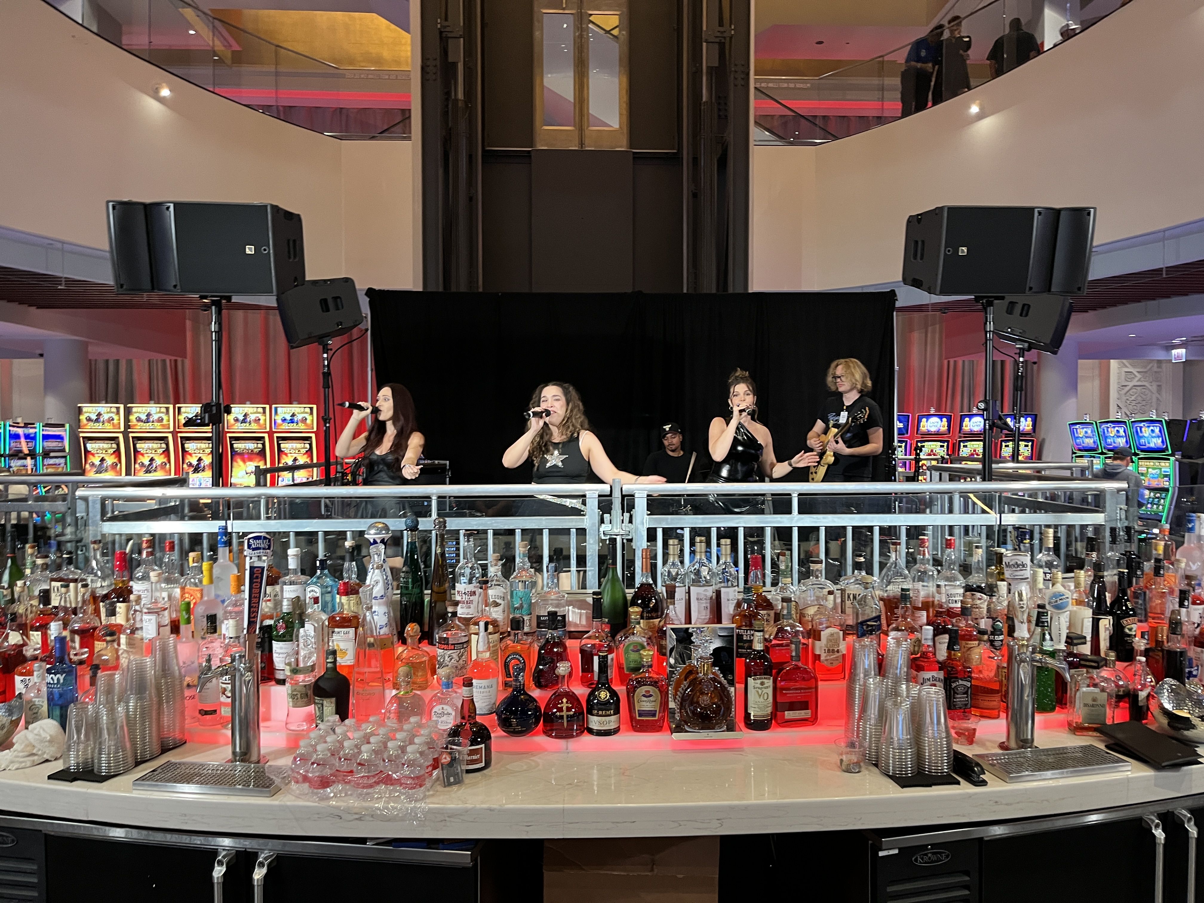 Band with three female singers and one male guitarist performing behind a bar filled with assorted liquor bottles and plastic cups in a brightly lit indoor venue with slot machines in the background.