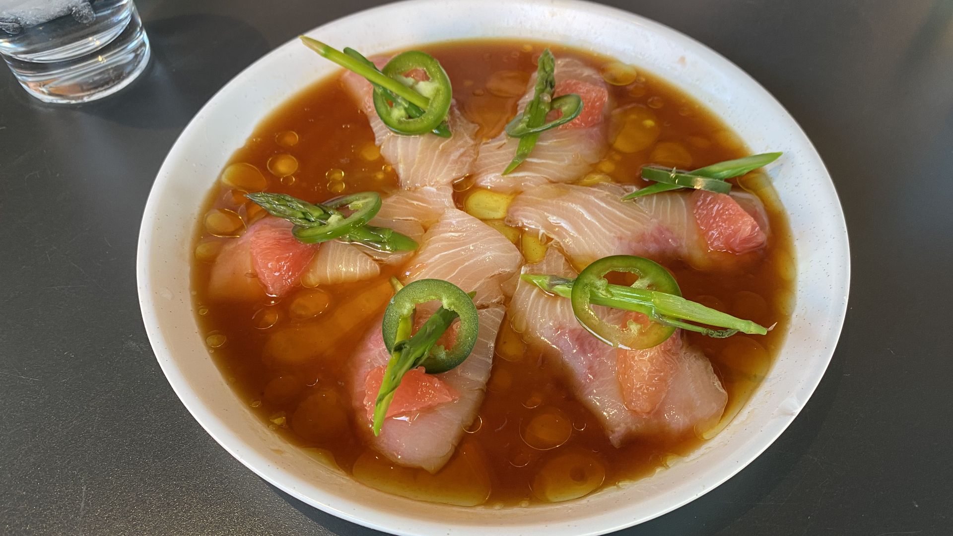 Lightly pink colored pieces of sashimi on a round plate with red sauce and topped with pieces of asparagus and jalapeno.