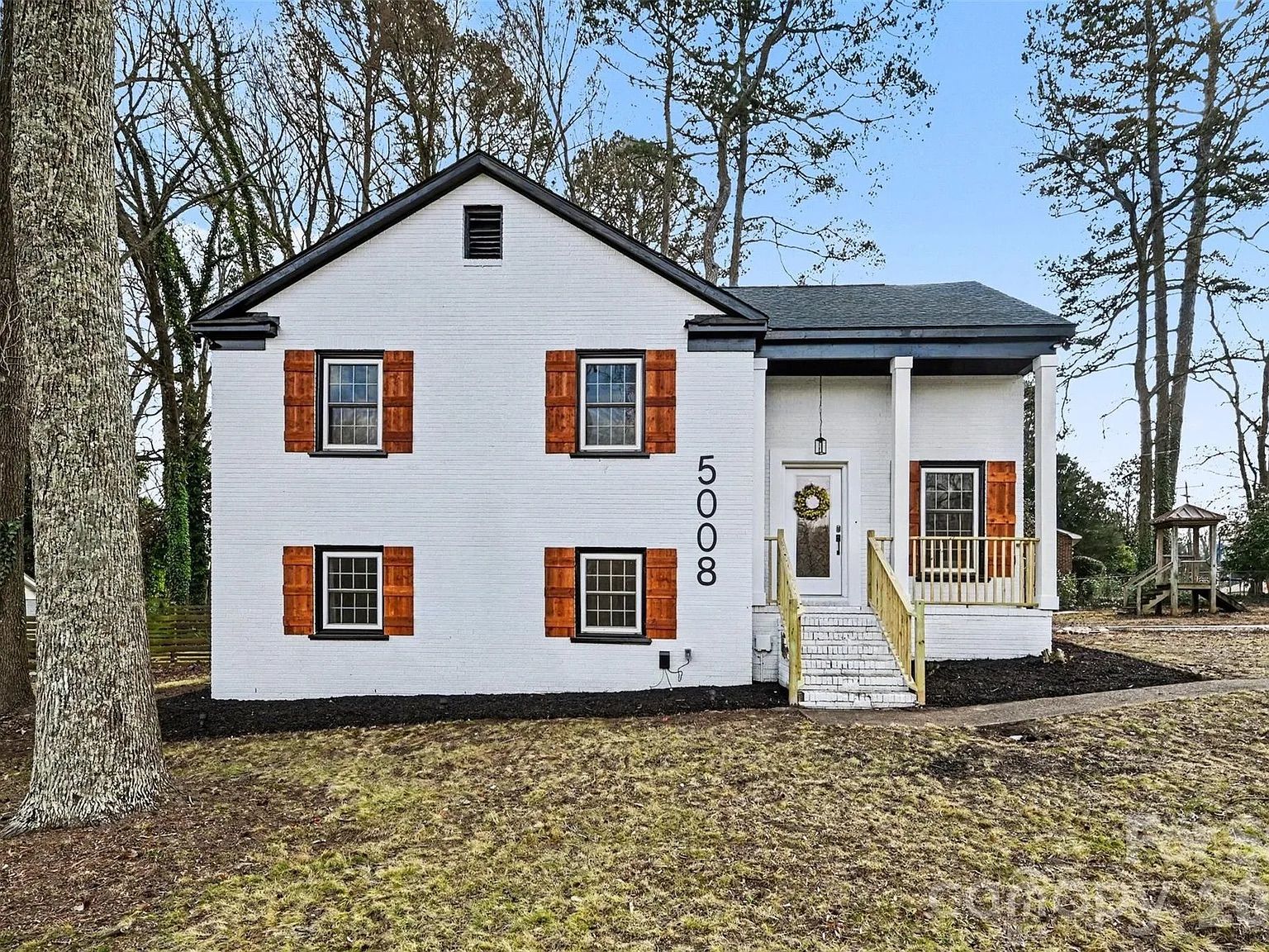 White brick house with black trim and wooden shutters, a front porch with stairs and railing, a wreath on the front door, sparse brown grass, tall leafless trees in background under clear blue sky.