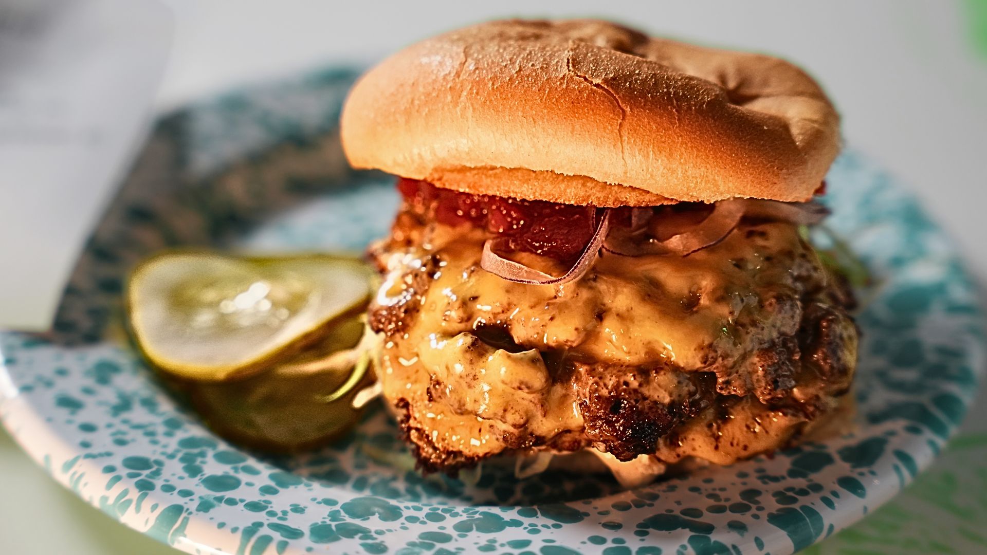 Cheeseburger with melted cheese, bacon, onions, and a toasted bun on a blue-speckled plate with pickle slices on the side.