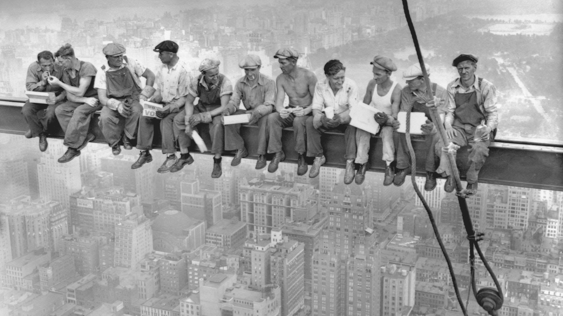 A  bunch of young workers sit on a ledge high above a city. its an old photograph in black and white and the men are wearing caps and overalls